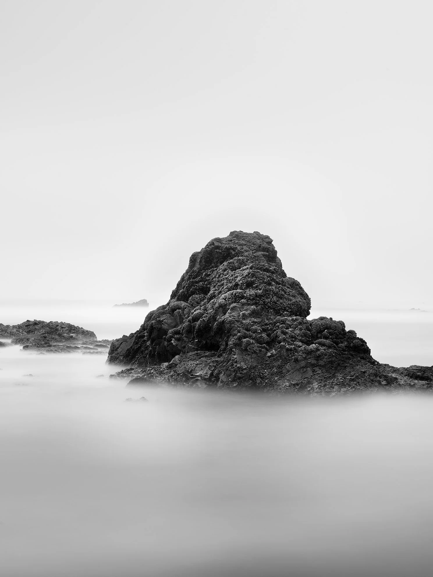 Black and white photo of a large rock formation in the ocean with smaller rocks around it, and a cloudy sky in the background.