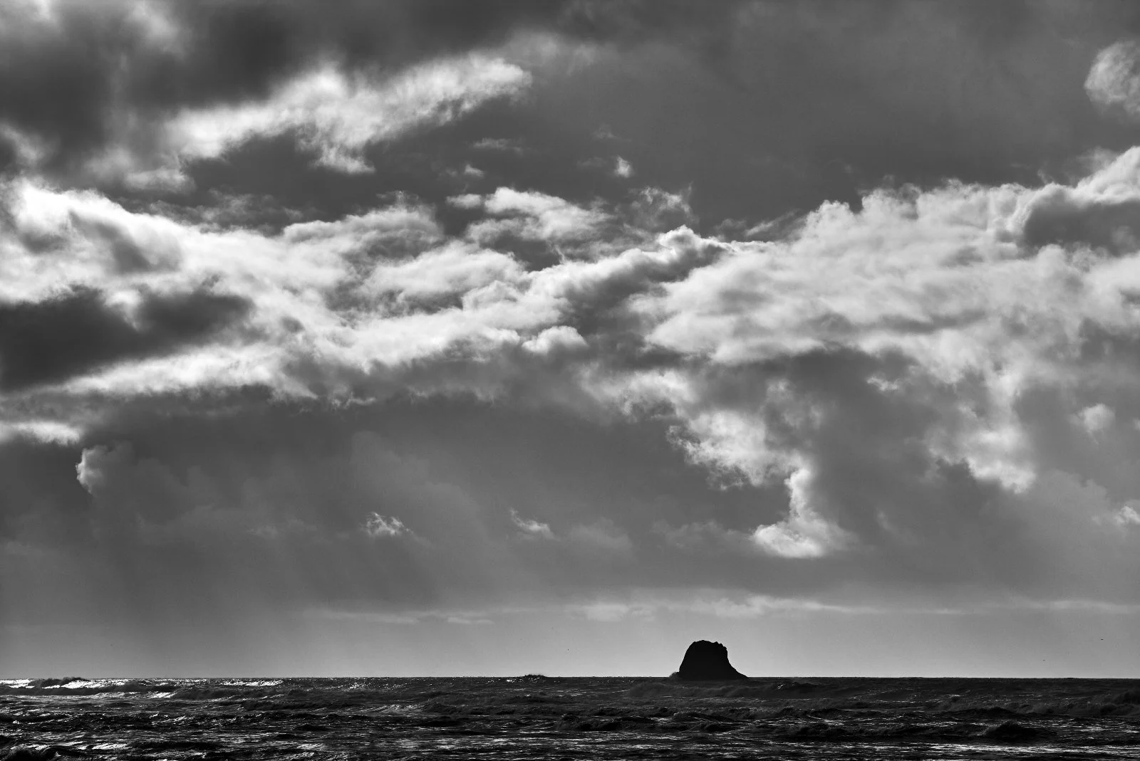 A black and white photograph of a seascape with a large rock formation in the distance. The sky is filled with dark, heavy clouds, creating a dramatic atmosphere.
