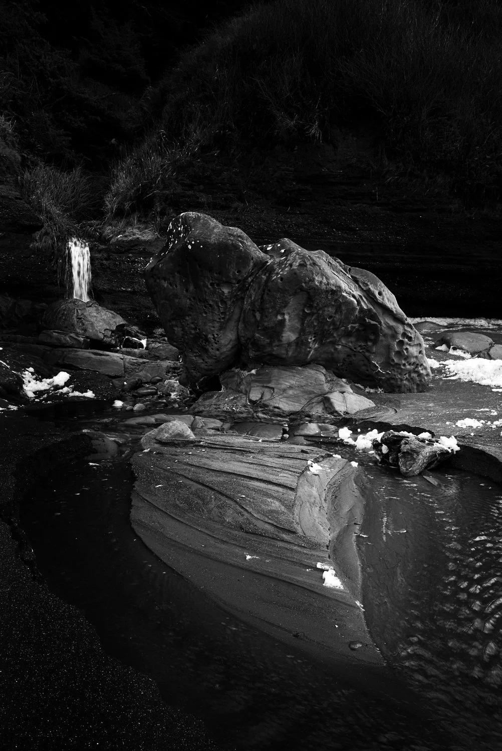 Black and white photo of a waterfall flowing over rocks into a small pool, surrounded by dark terrain and vegetation.