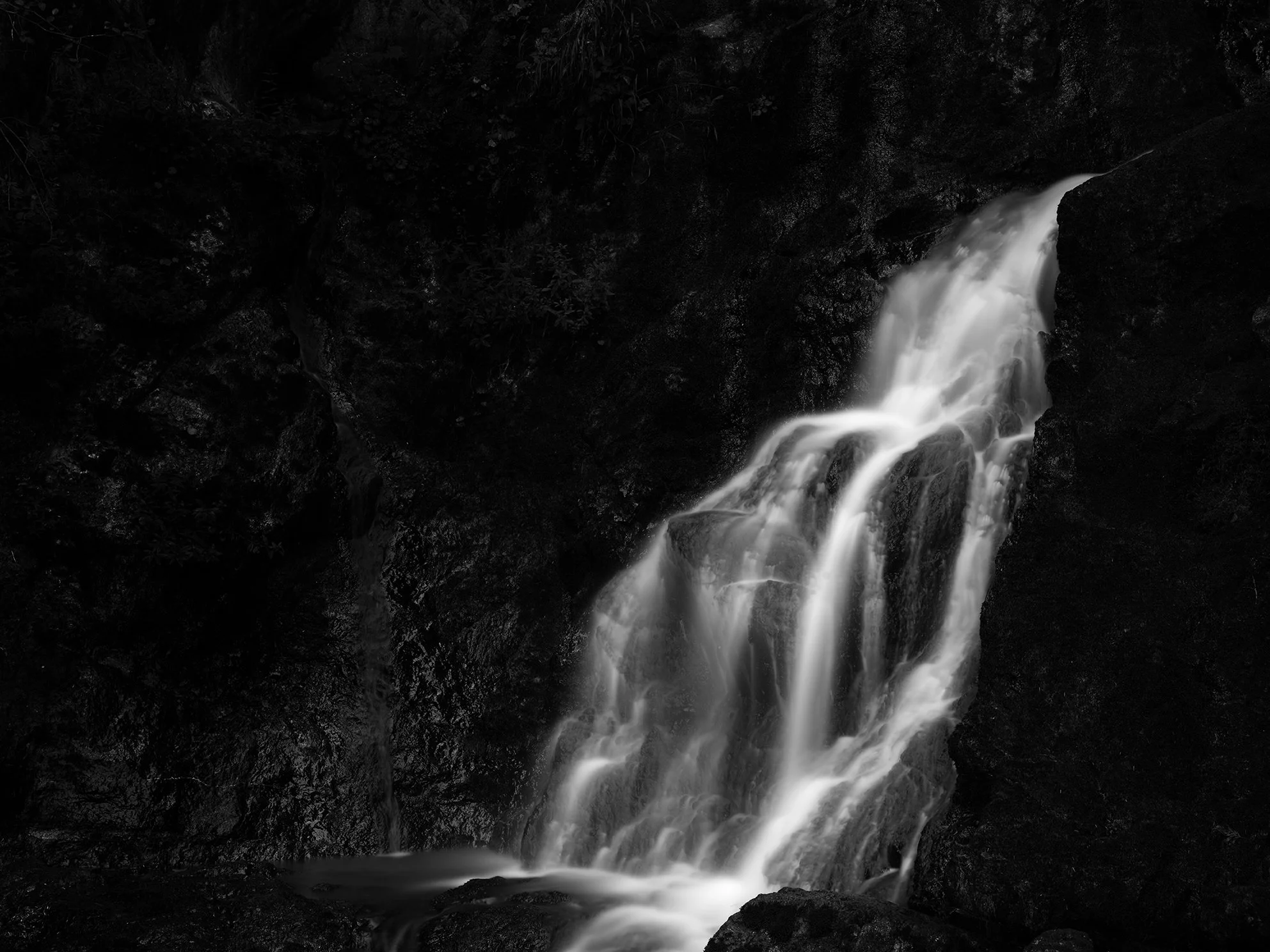 A black and white photograph of a waterfall cascading down rocks in a dark, forested area.