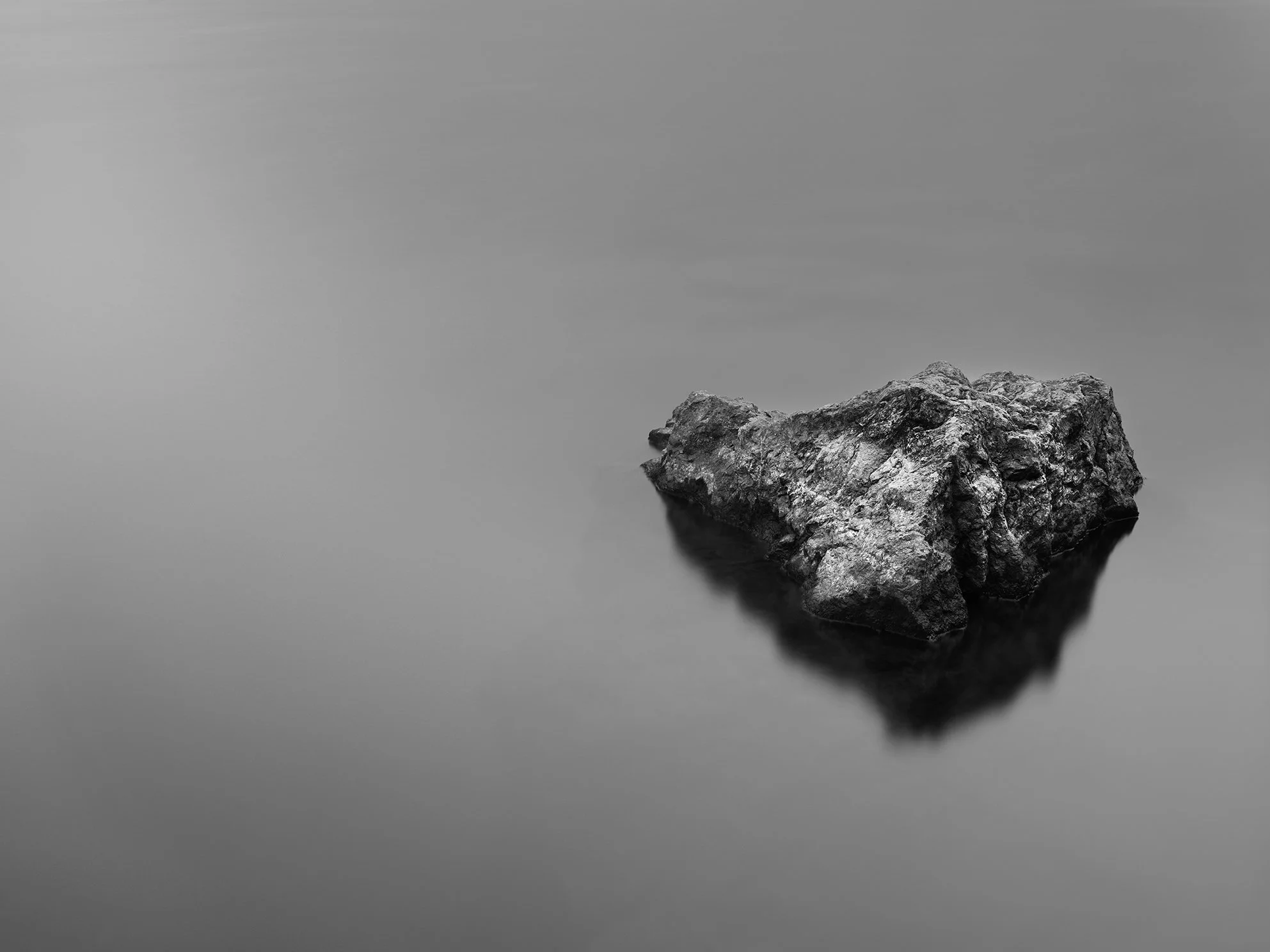A large rock resting in calm water with a smooth, reflective surface, creating a mirror image of the rock and an overcast, gray sky.