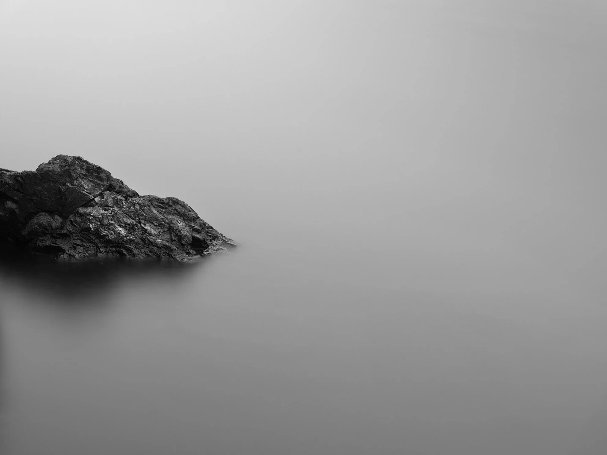 Black and white photograph of a rocky formation on the left side with misty water and a foggy sky.