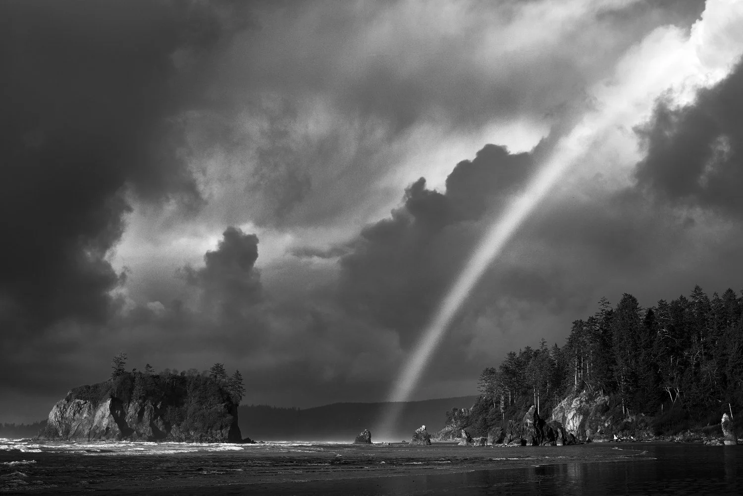 A black and white photo of a dramatic sky over a rugged coastline, featuring a rainbow piercing through the clouds and touching the water, with trees on the shoreline.