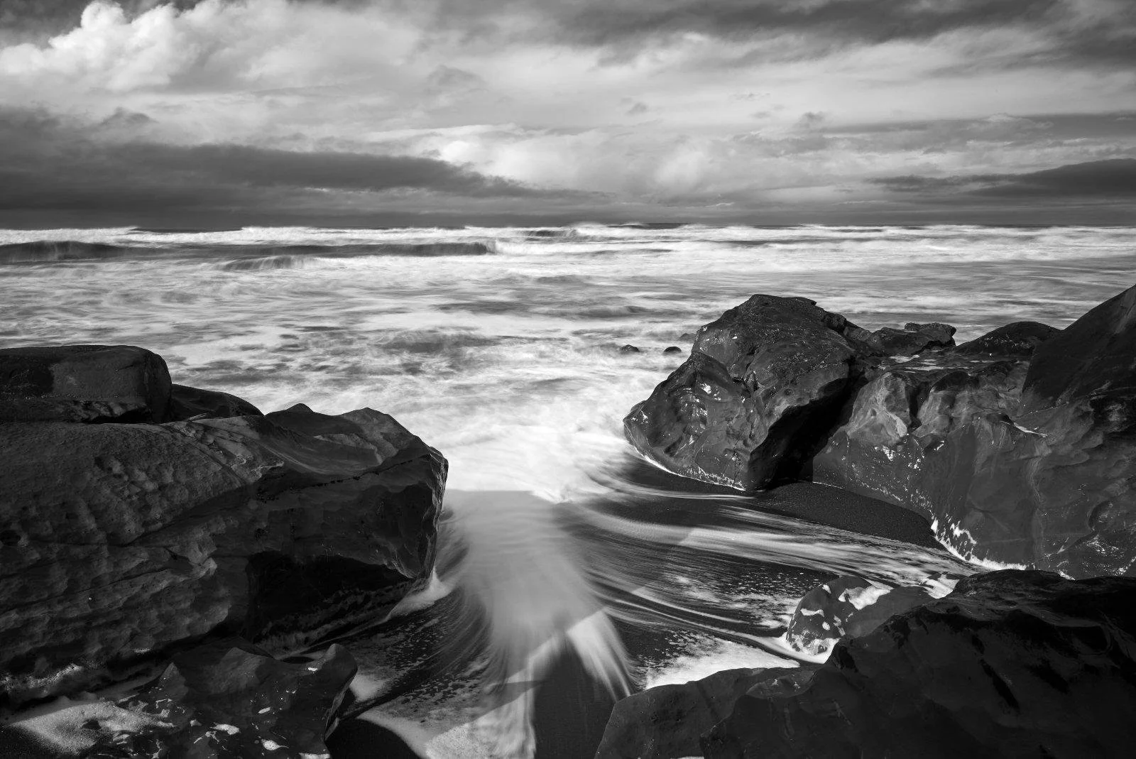 Black and white photograph of a rocky shoreline with waves crashing and swirling water, under a cloudy sky.