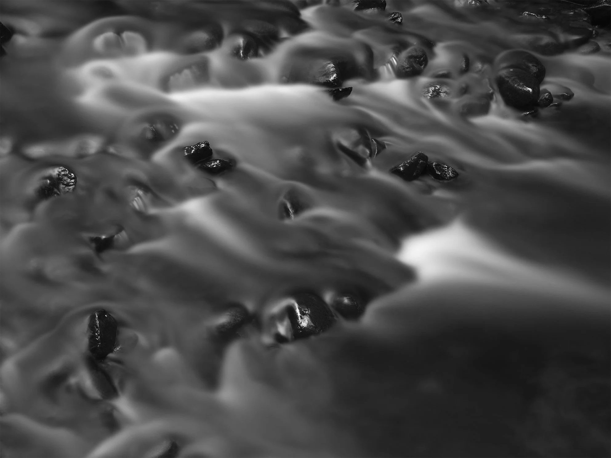 Close-up of water flowing over small dark stones in a river, in black and white.