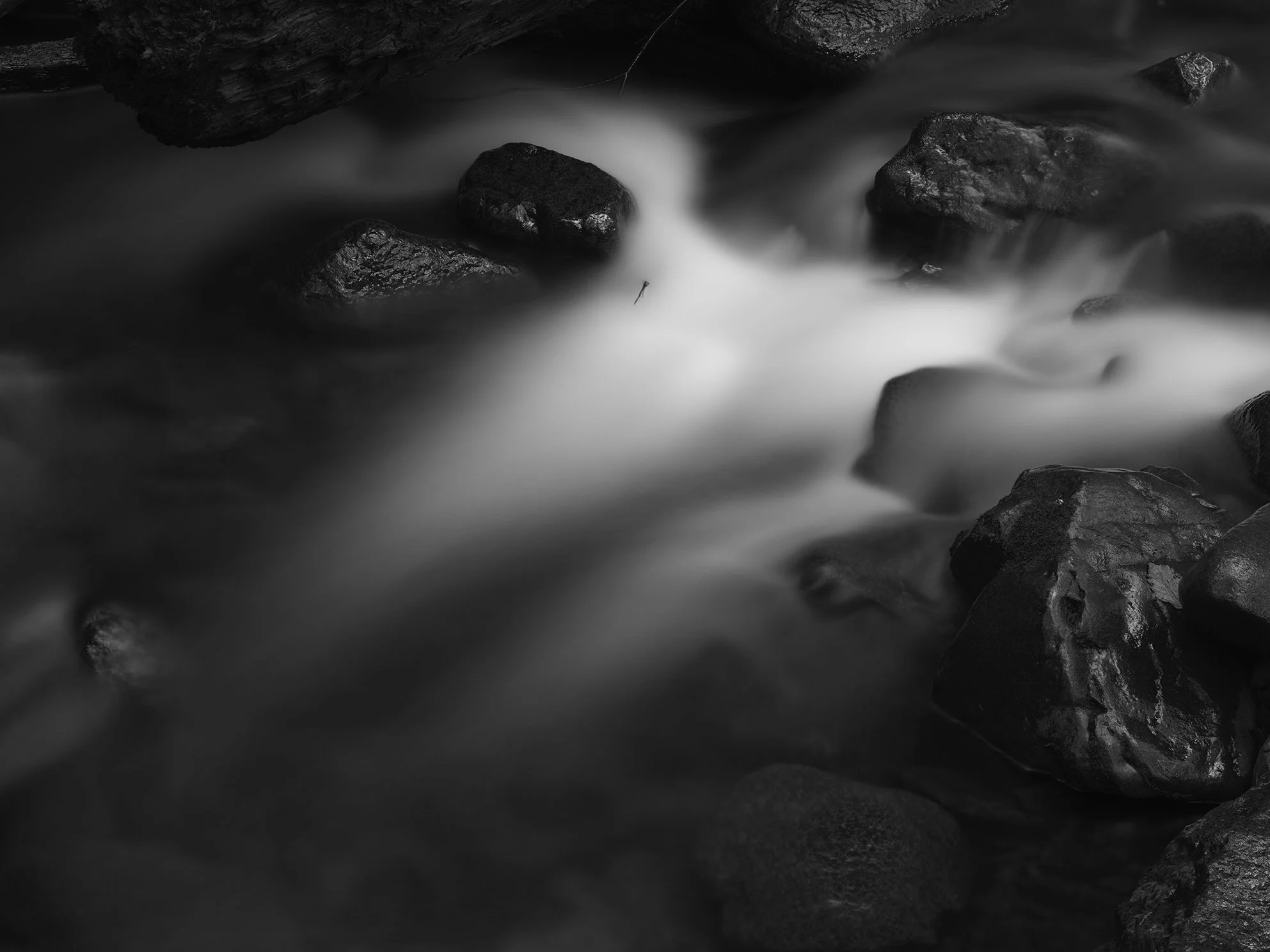 Black and white photograph of a rocky stream with water flowing over rocks, appearing smooth due to long exposure.