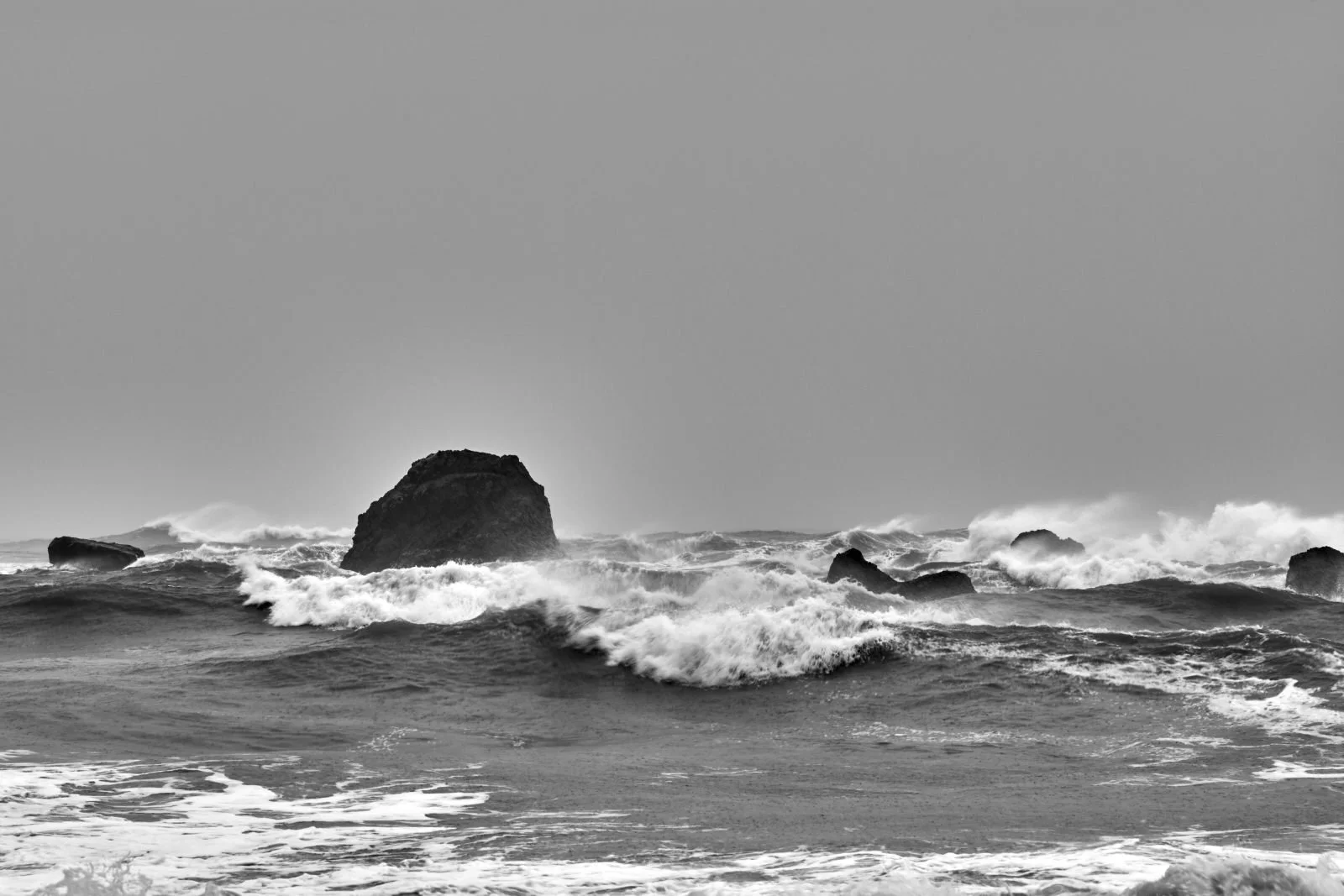 Black and white photo of ocean waves crashing around large rocks and boulders near the shoreline under a clear sky.