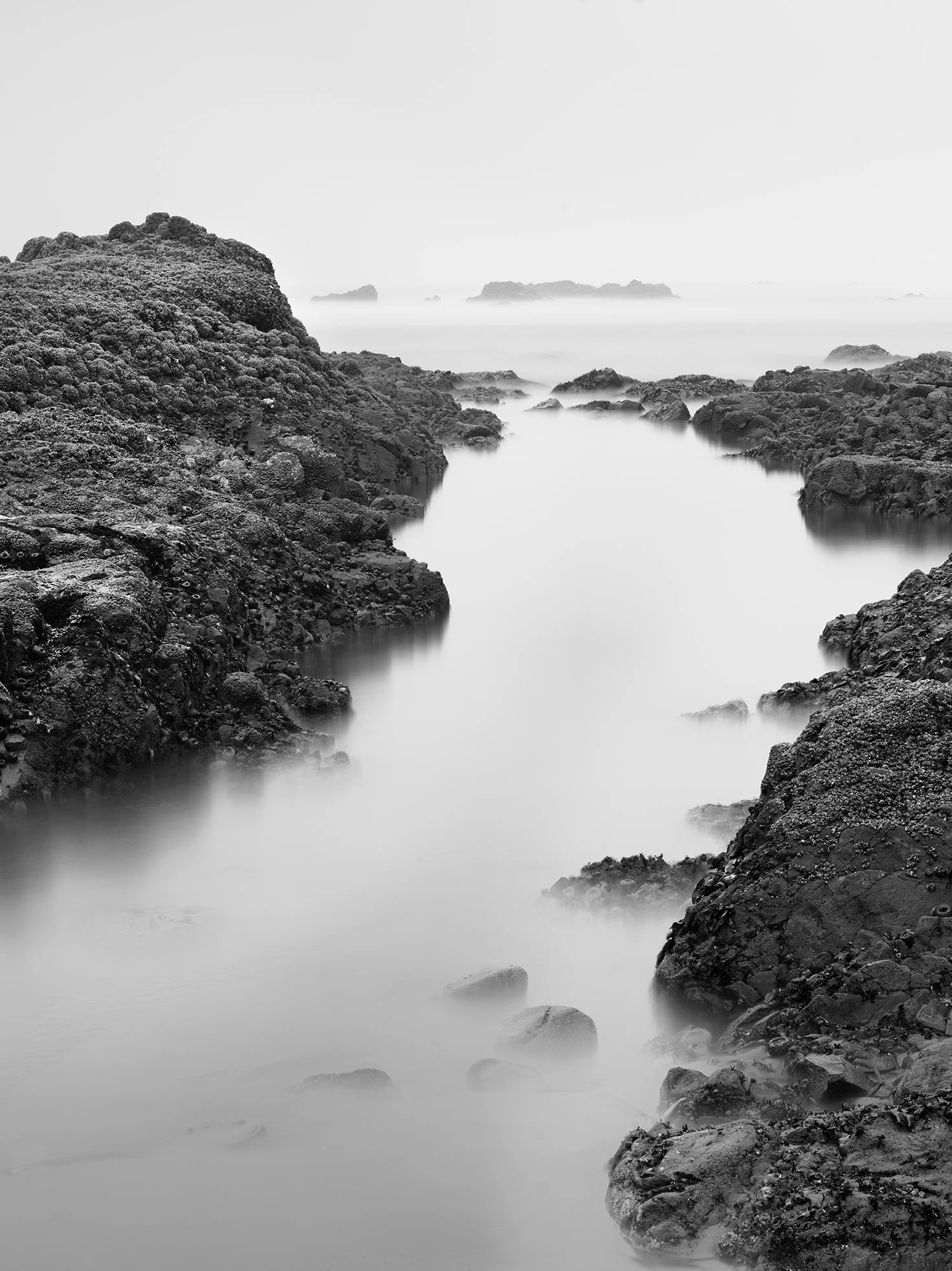 Black and white long-exposure photograph of rocky coastal landscape with calm water and distant islands.