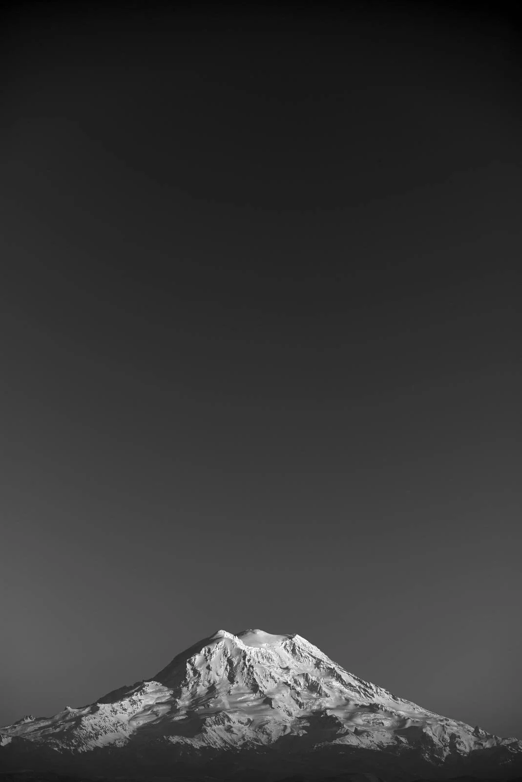 A black and white photograph of a snow-capped mountain, likely Mount Hood, with a dark sky above.