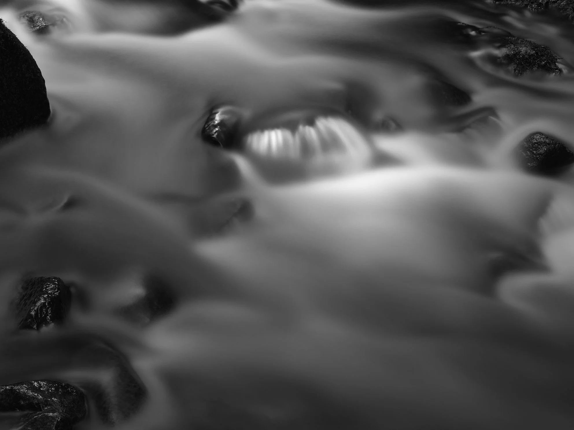 Black and white long exposure photograph of a flowing river with visible rocks submerged in the water.