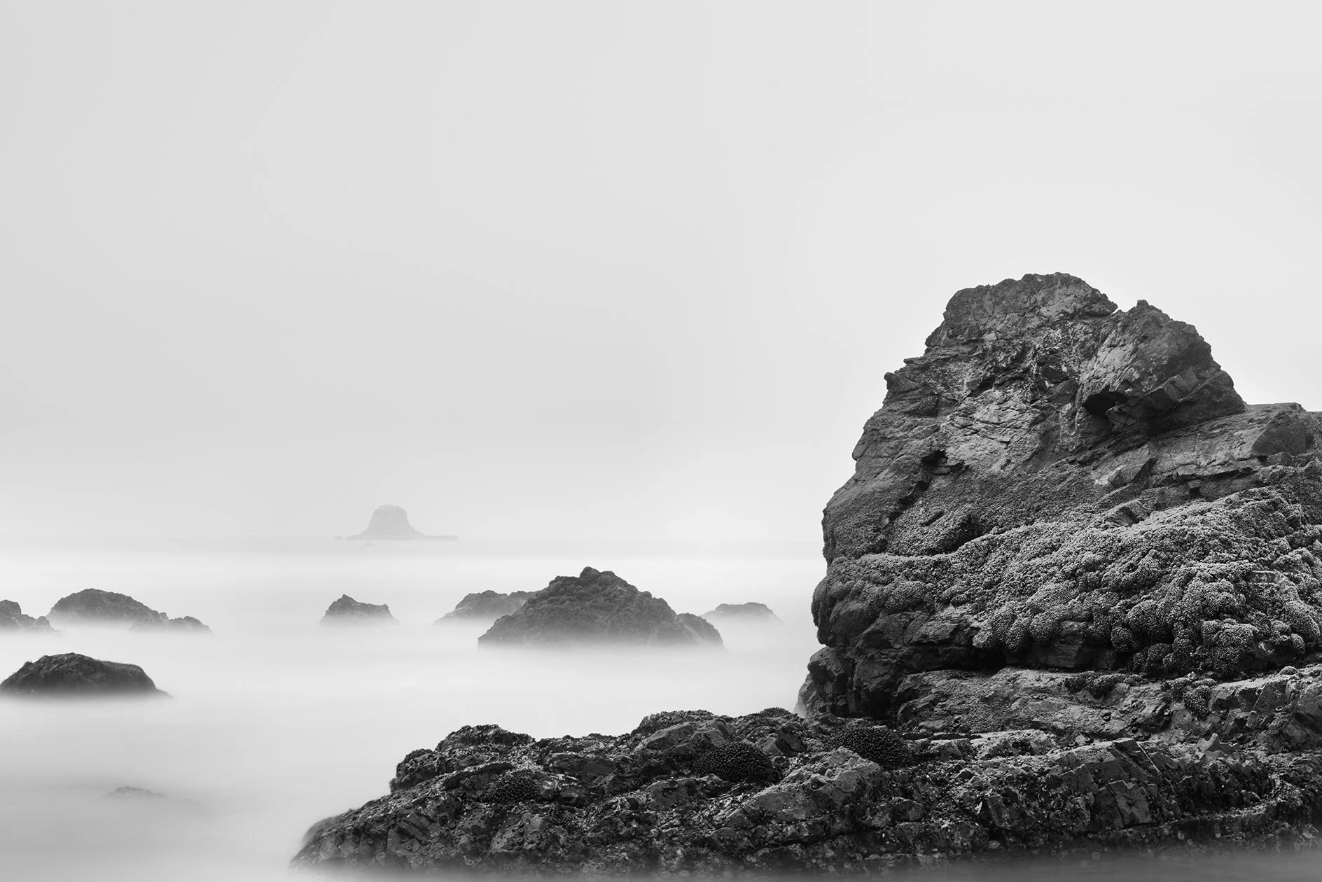 Black and white photo of rocky shoreline with large rocks near the water and a small rock formation in the distance under overcast sky.