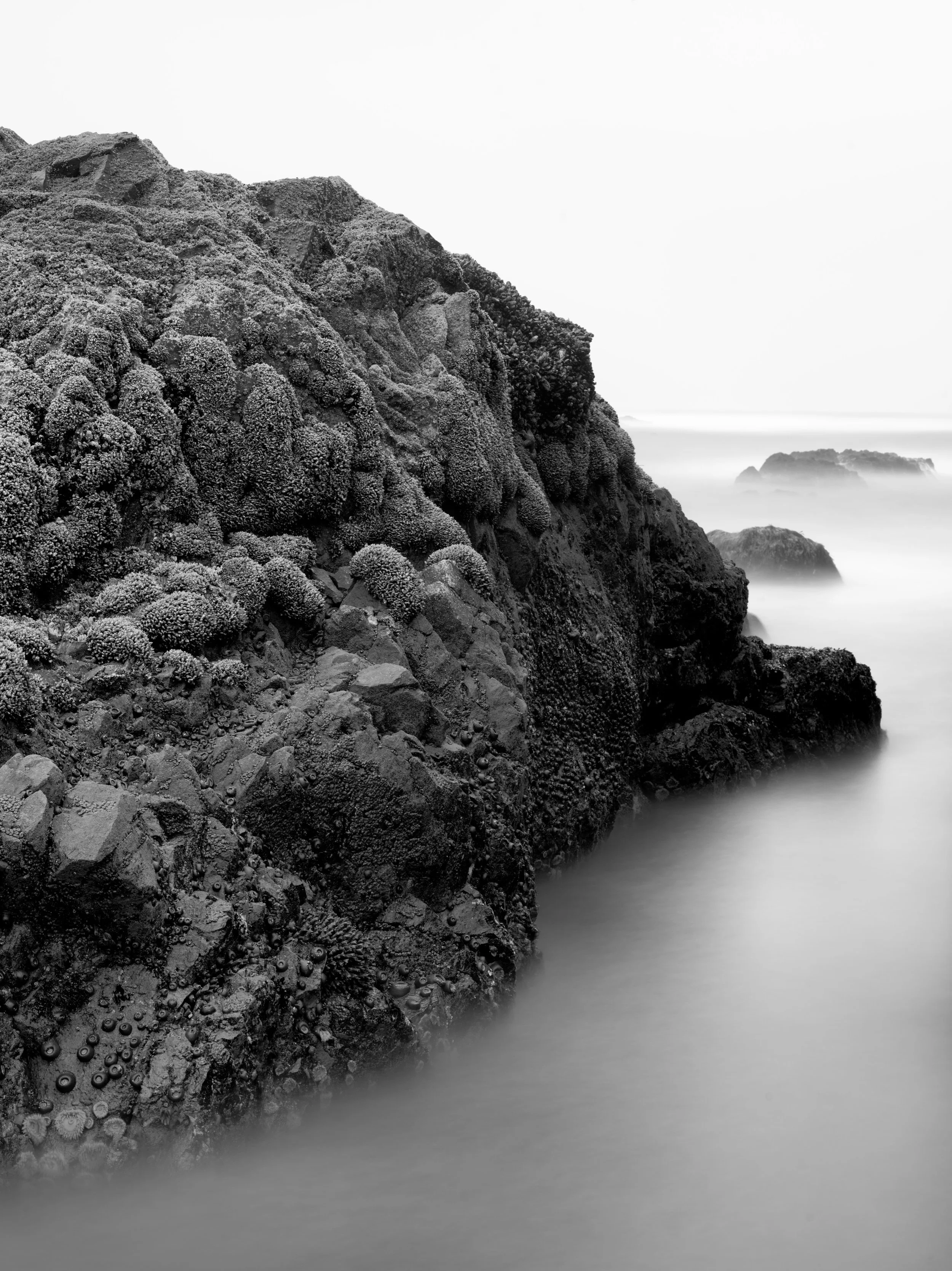 Black and white photo of rugged seaside cliffs with textured rocks, water seemingly smooth and misty in the foreground, and distant rocky islands or formations on the horizon.