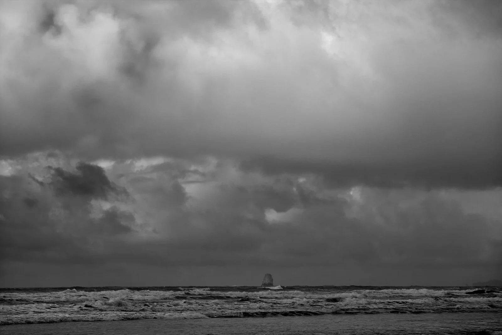 Black and white photograph of a cloudy sky over the ocean with waves and a large rock formation in the distance.