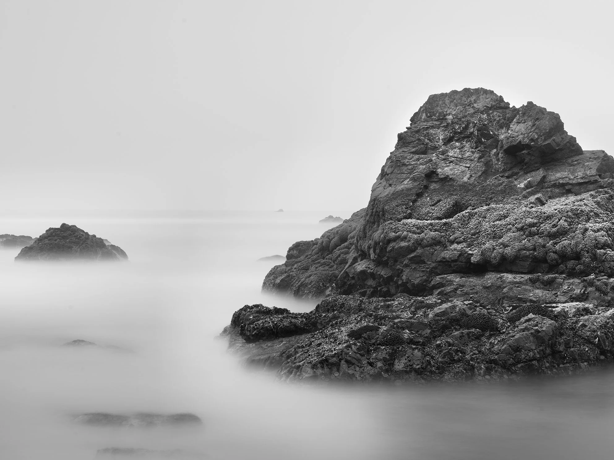 A black and white photo of dark, rugged rocks along a coastline with mist or long exposure water surrounding them, creating a surreal, calm atmosphere.