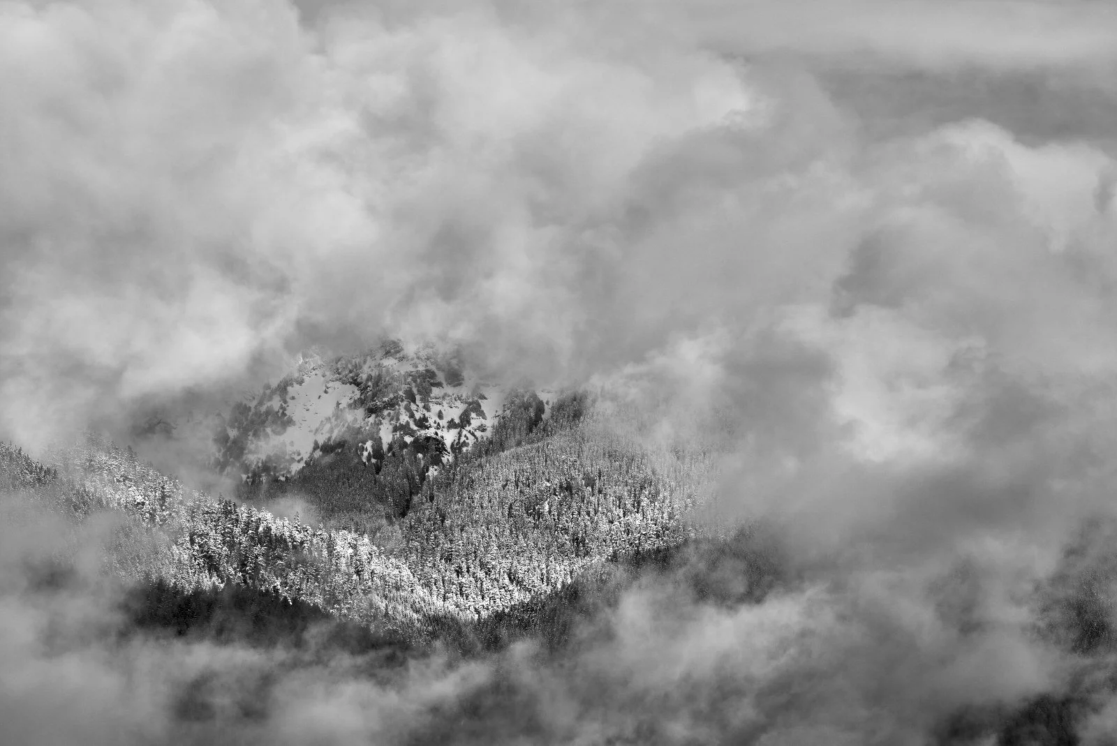 Black and white photograph of snow-covered mountain peaks surrounded by clouds and mist.
