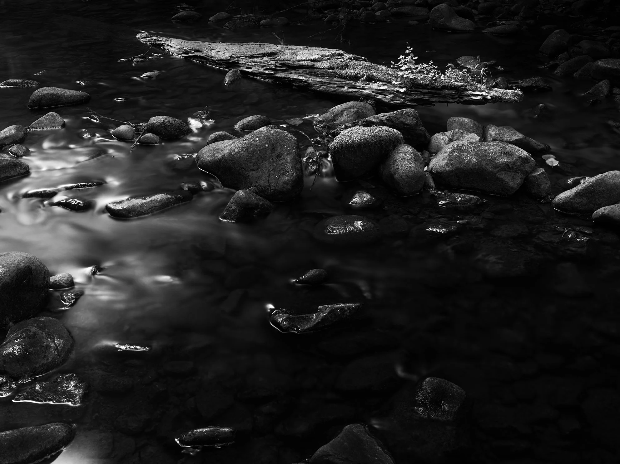 A black-and-white photo of a rocky stream with smooth stones and a fallen log in the water.