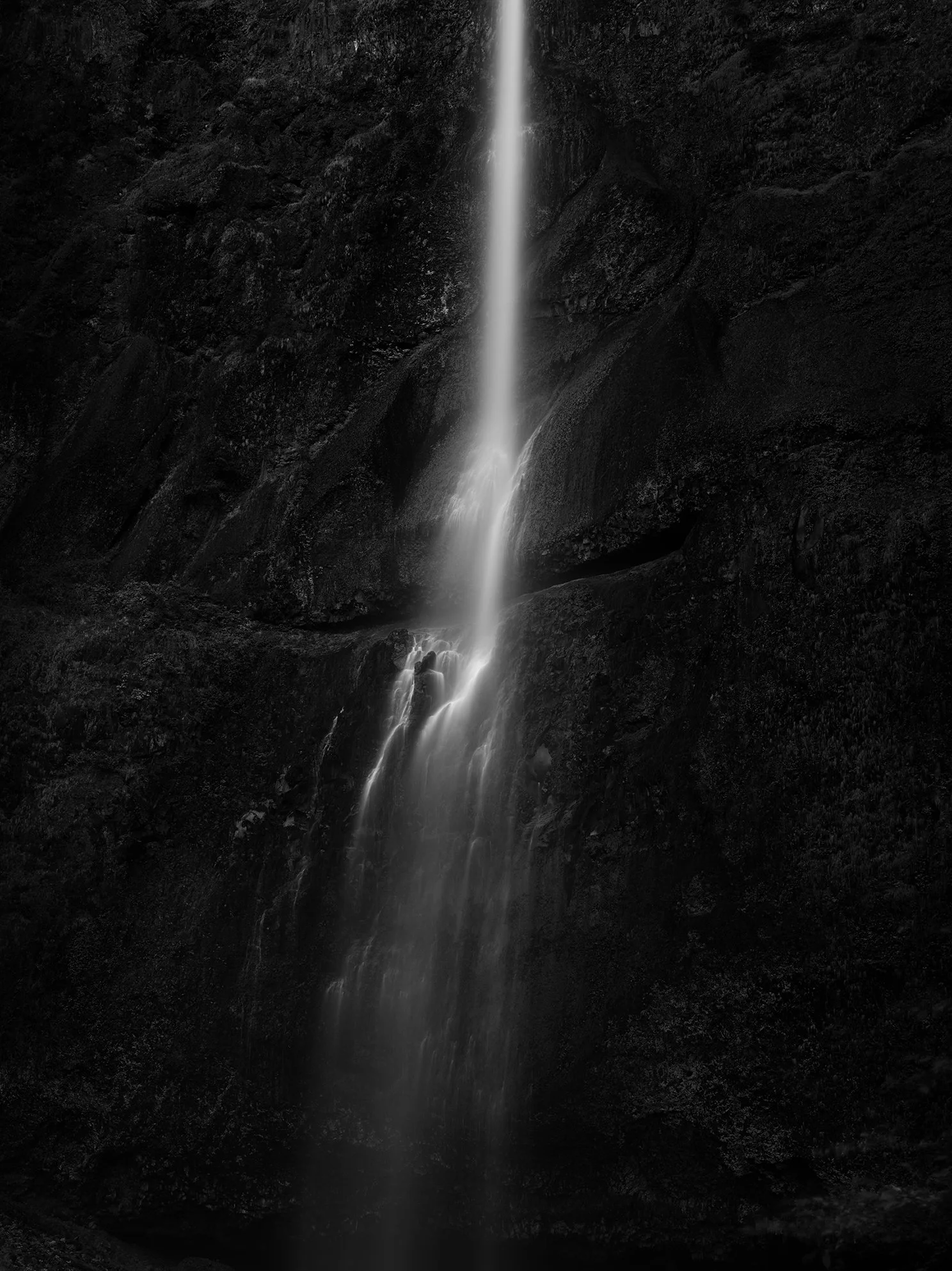 A narrow waterfall flowing down dark, rocky cliffs in a black-and-white photograph.