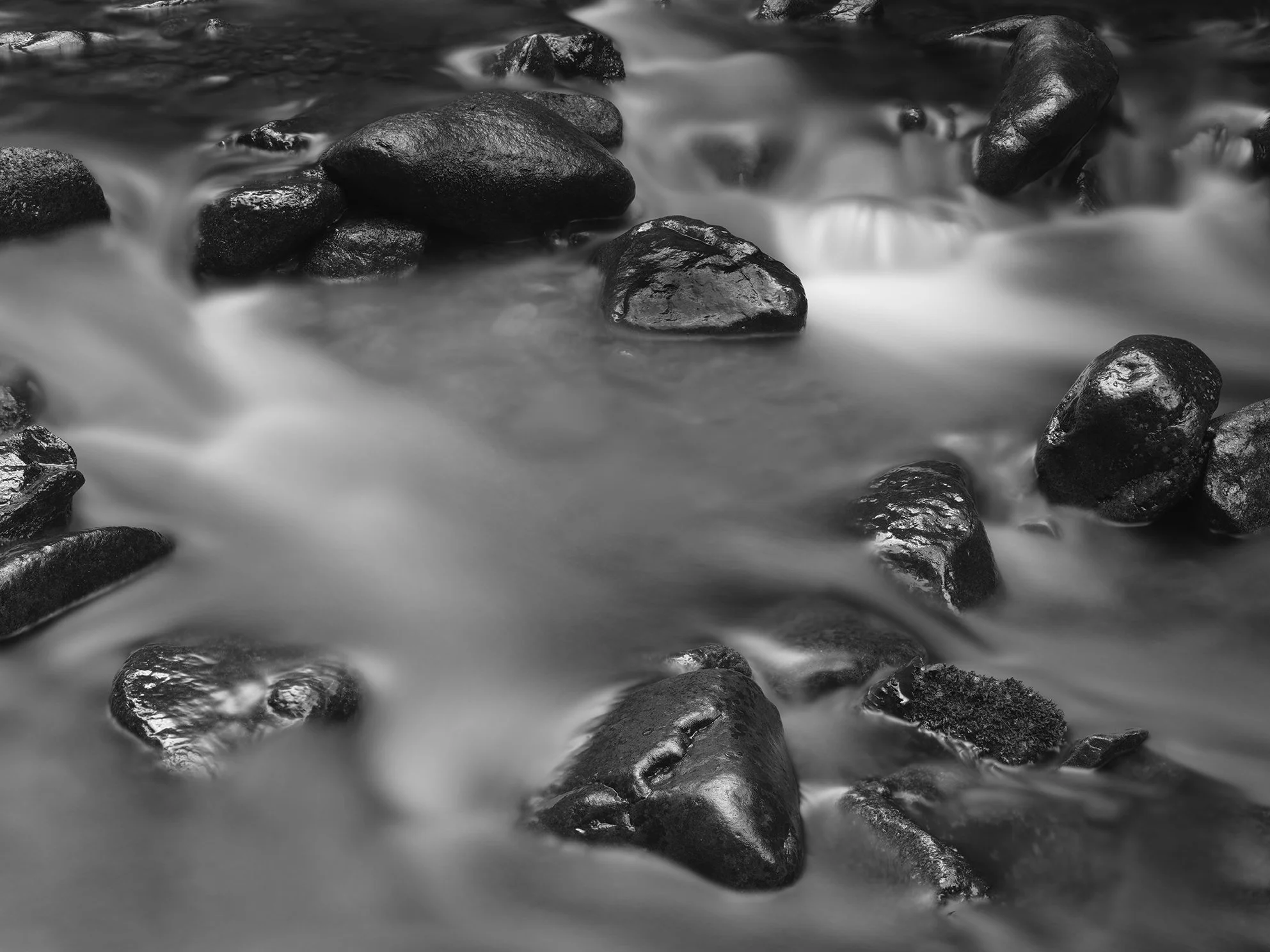 Black and white photograph of smooth water flowing around rounded rocks in a stream.