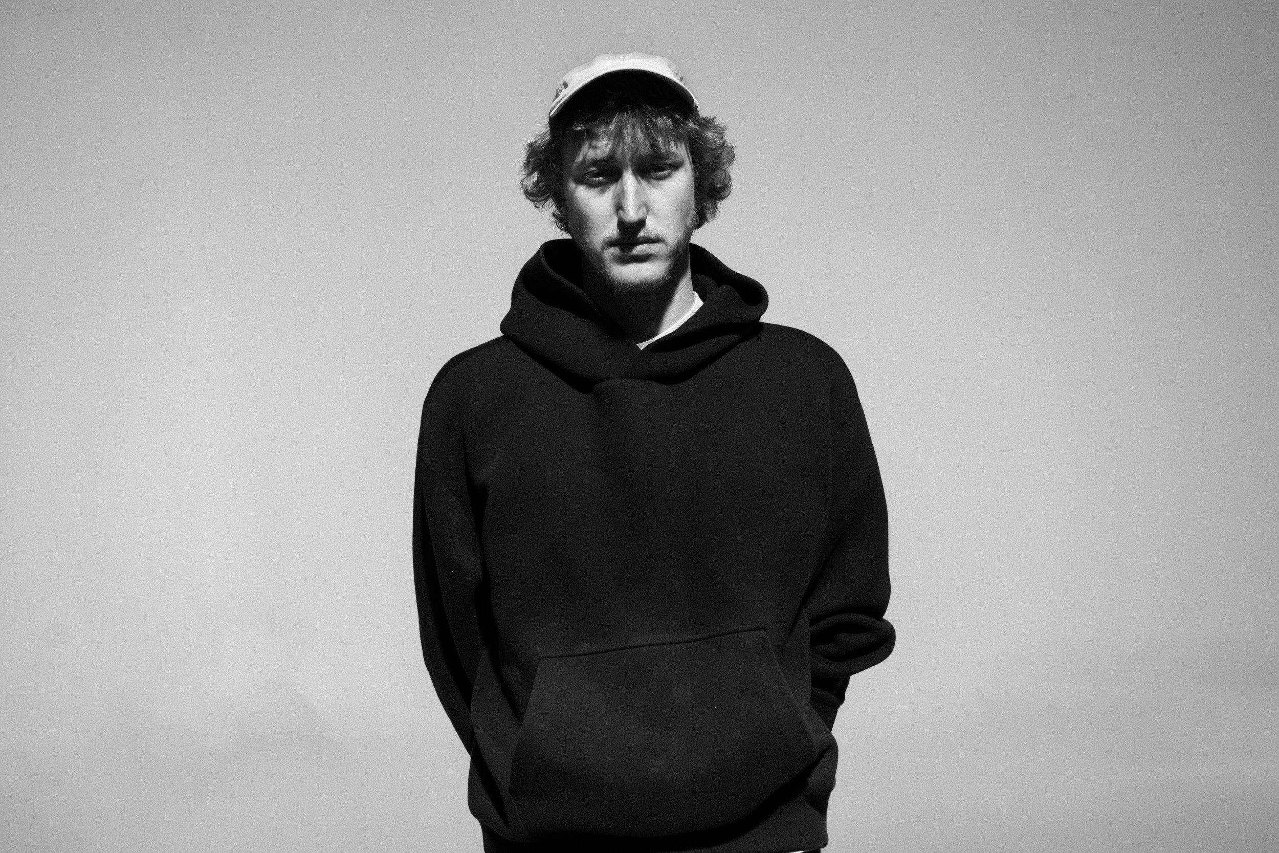 Black and white portrait of a person wearing a hoodie and cap, looking at the camera.