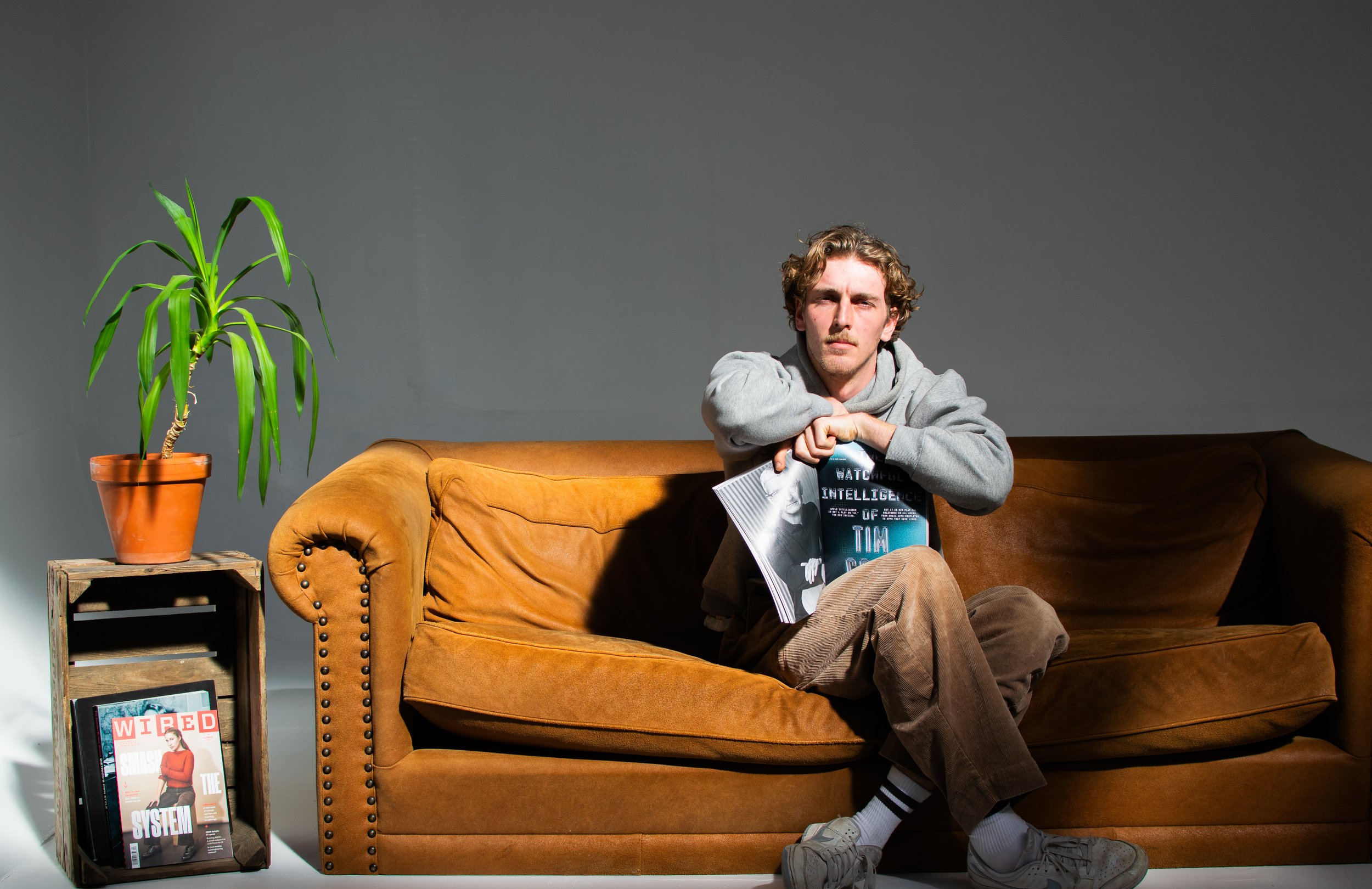 Man sitting on a brown sofa holding a magazine, with a potted plant and a wooden crate holding a magazine next to him in a photostudio