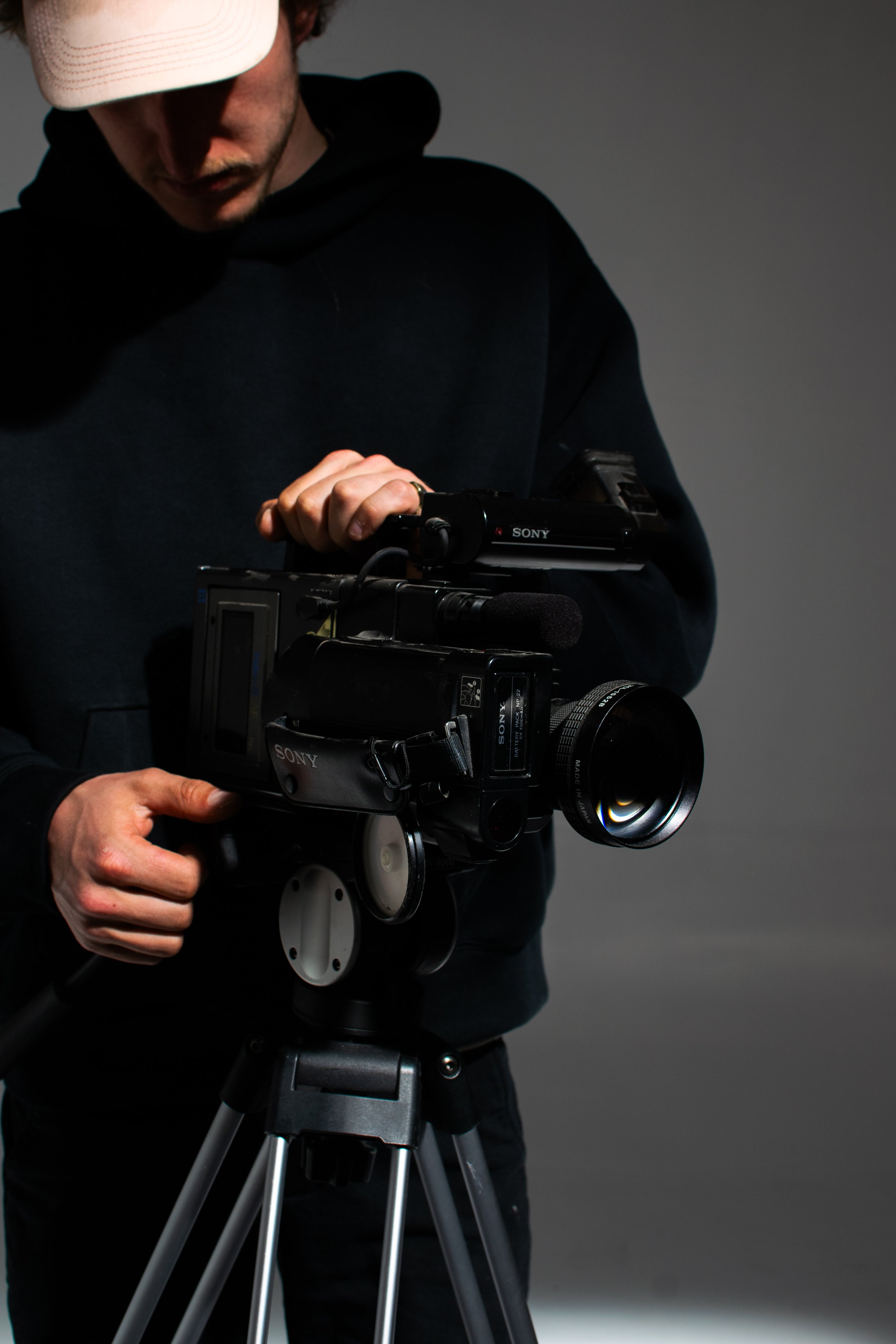 Person operating a Sony video camera on a tripod, wearing a dark hoodie and a light cap.