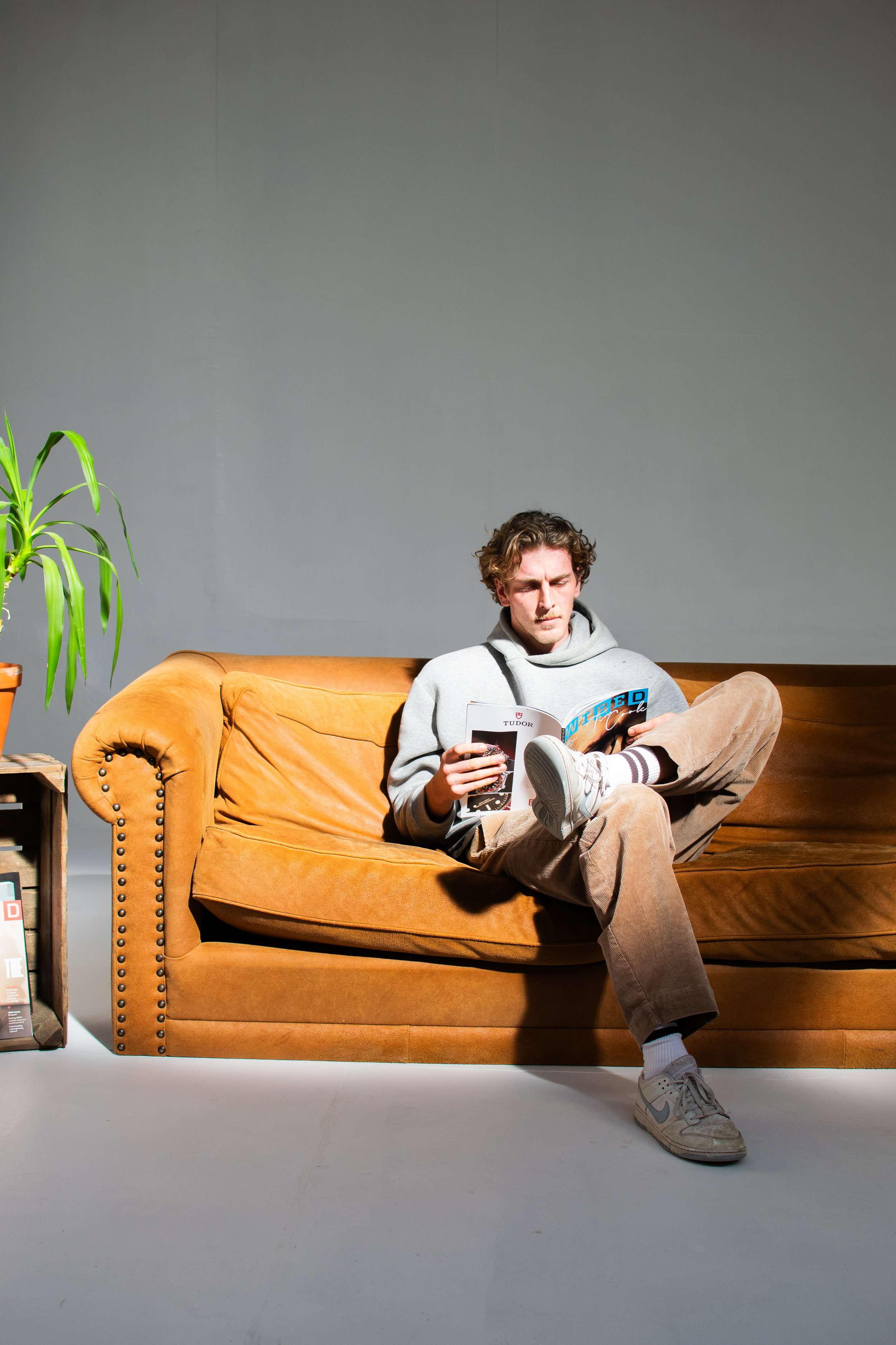 Man sitting on a brown sofa reading a magazine, with a green plant on a side table in a photostudio