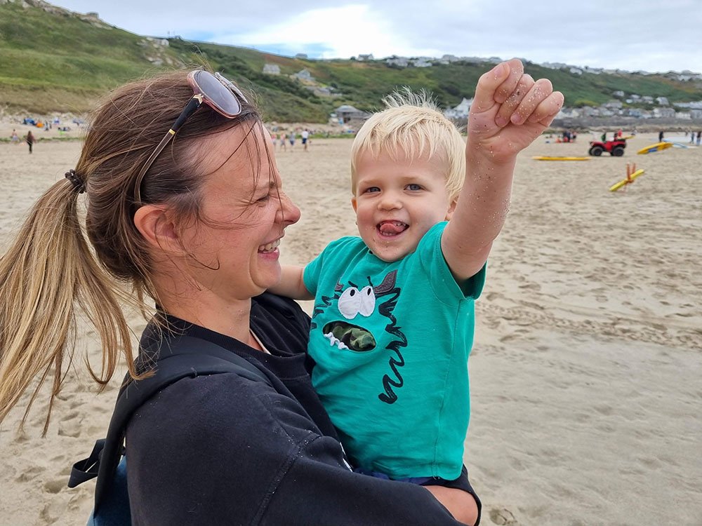 A woman and a young boy at the beach, with the woman smiling and the boy raising his fist in excitement, both enjoying a day in the sand with hills and cloudy sky in the background.
