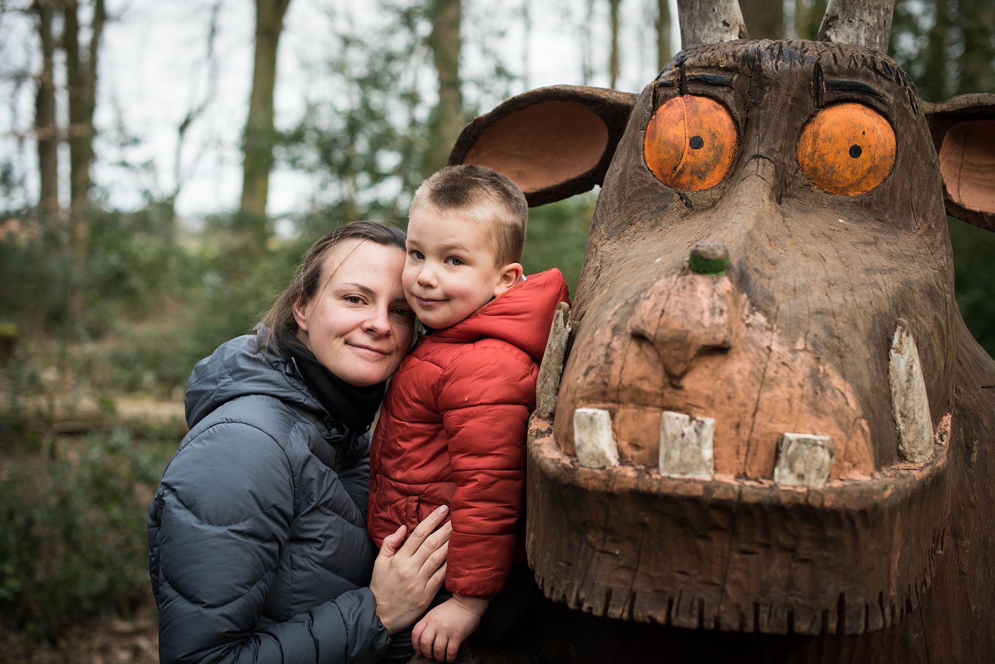 A woman and a young boy pose next to a large wooden totem carving of a crocodile with orange eyes and tusks in a forest setting.