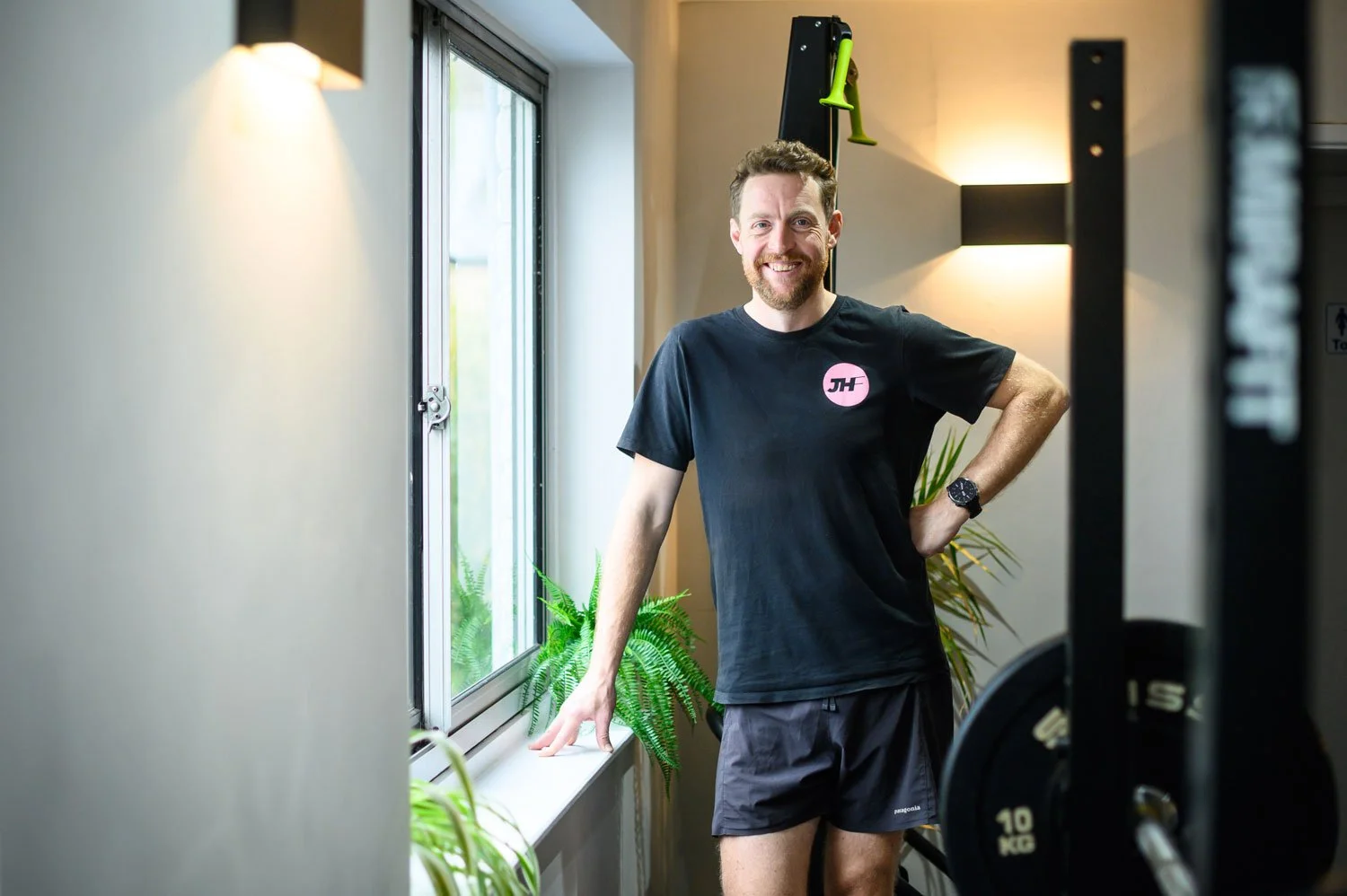 A man with a beard and short hair smiling, wearing a black t-shirt with pink logo and black shorts, standing indoors next to a window with plants, pose with one hand on his hip and the other resting on the window sill.
