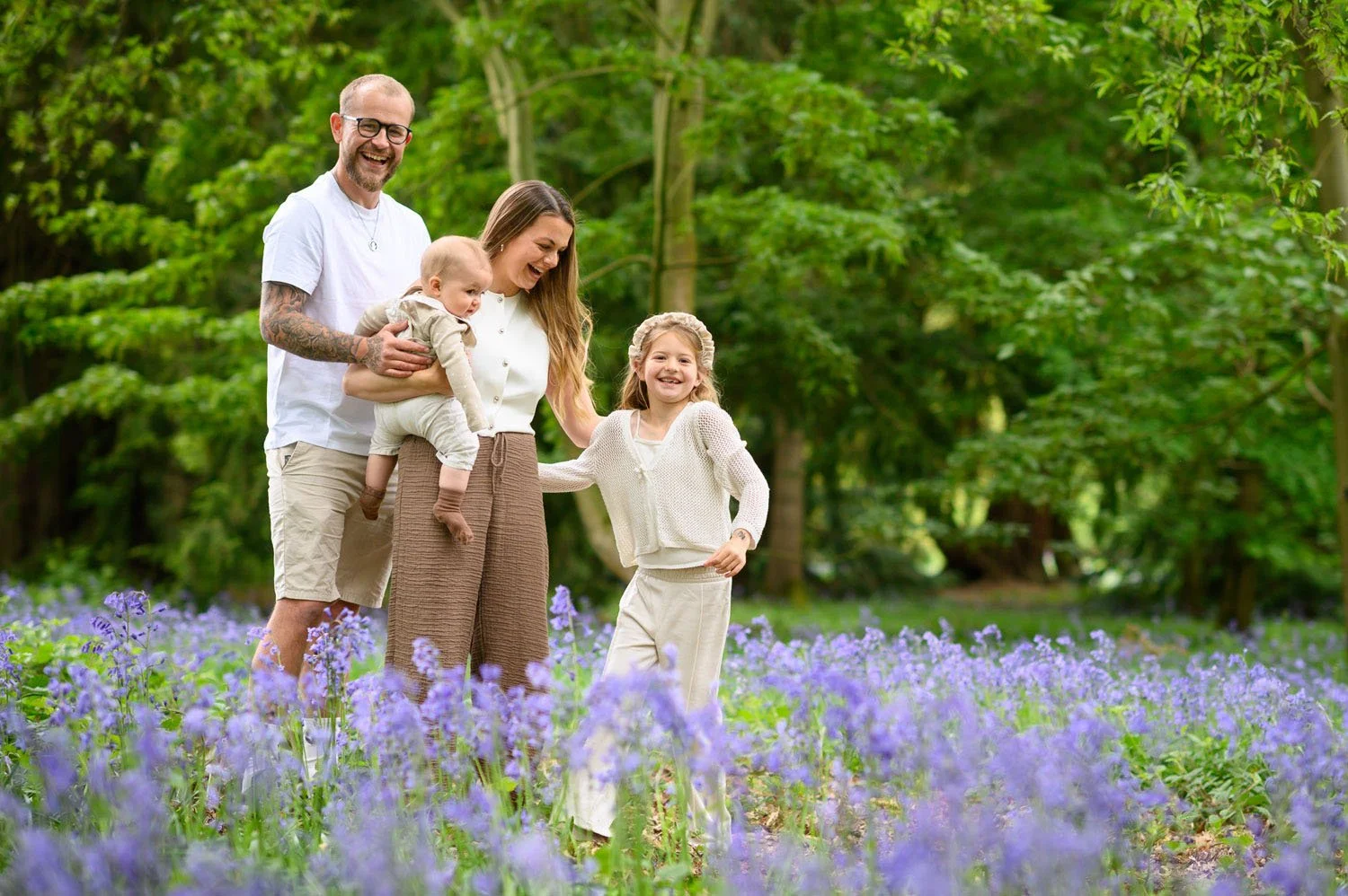 A happy family of four standing in a field of purple flowers in a lush green forest, smiling and enjoying the outdoors.