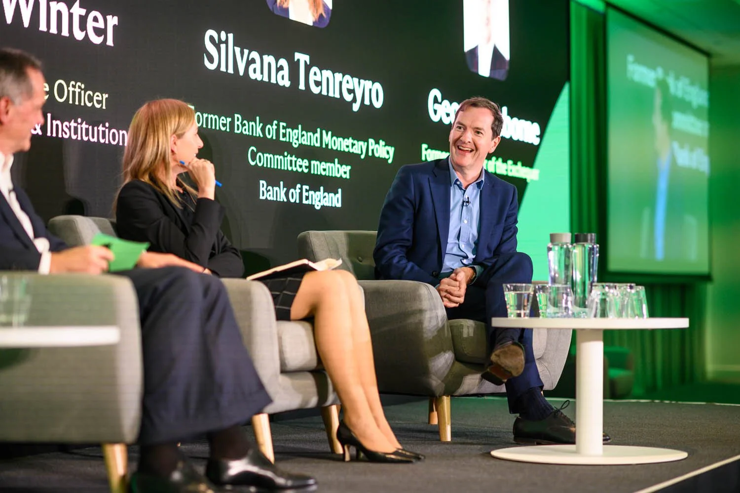 Three people sitting on stage with a large screen behind them displaying names and titles, engaged in a discussion at a formal event.