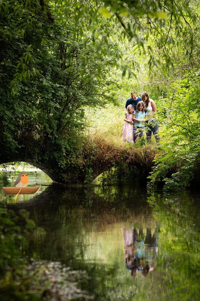 A family of four stands on a small grassy bank by a creek in a lush green forest. A cartoon illustration of a bird in a boat is reflected on the water.