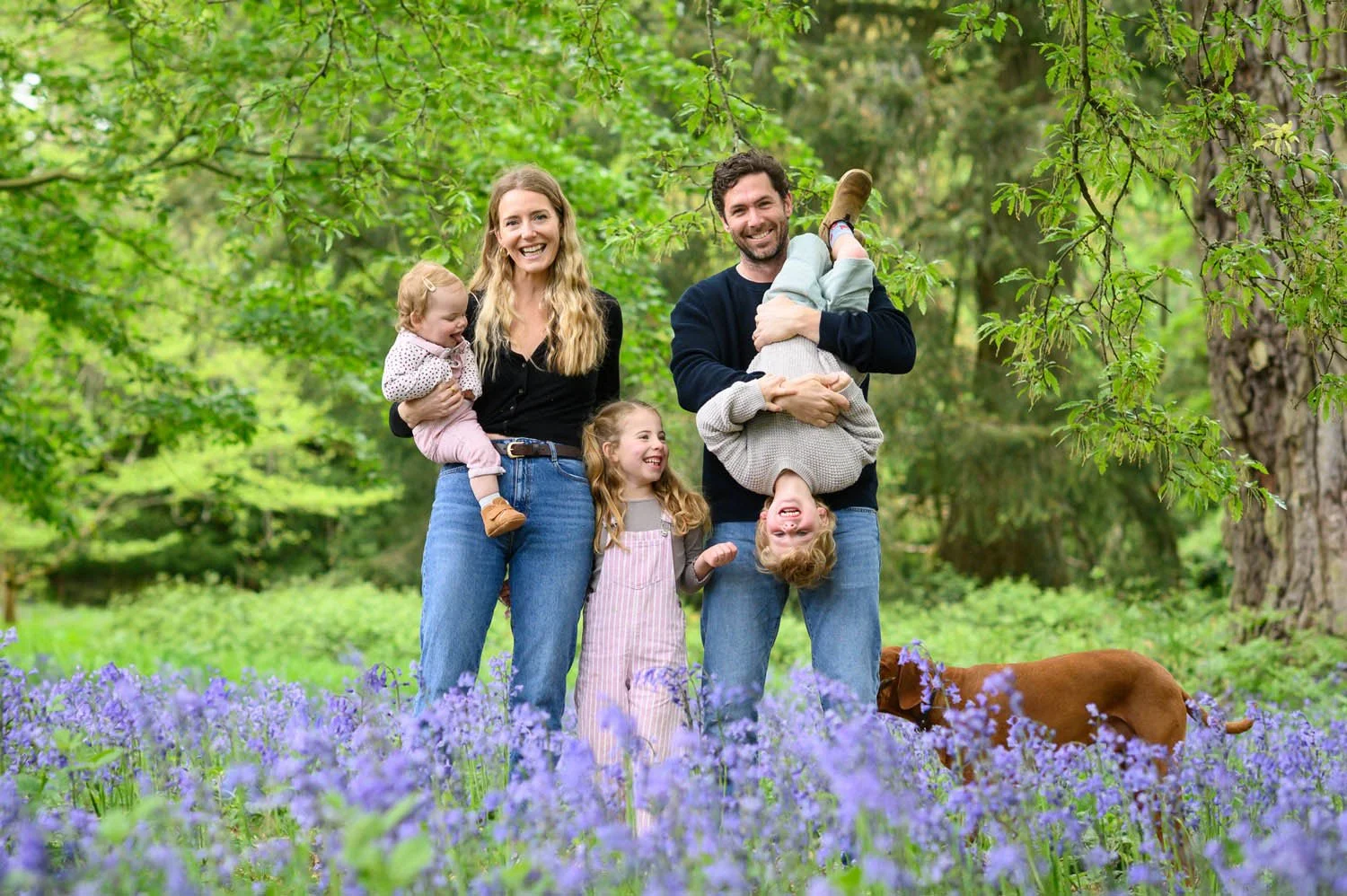 A family of five, including two adults and three children, standing in a lush green forest with purple flowers on the ground and a brown dog nearby. The adults are holding two of the children, and the children are smiling and laughing.