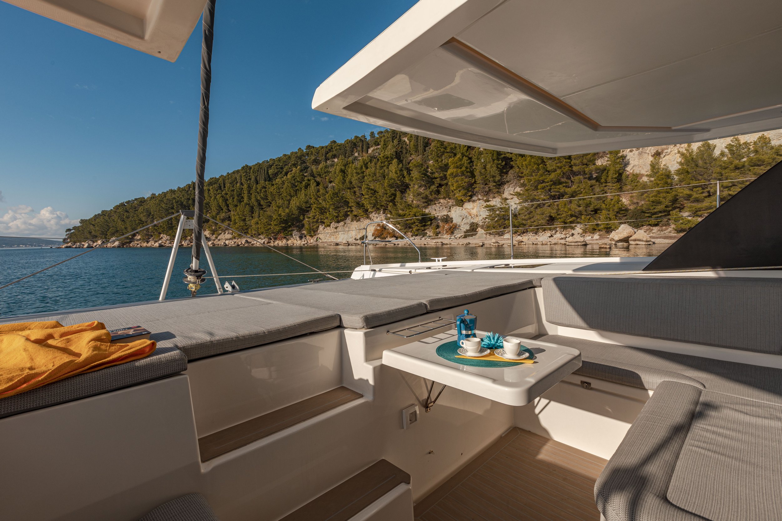 View from a yacht's outdoor seating area showing cushions, a table with tea cups, a lantern, and a scenic view of water and forested shoreline.