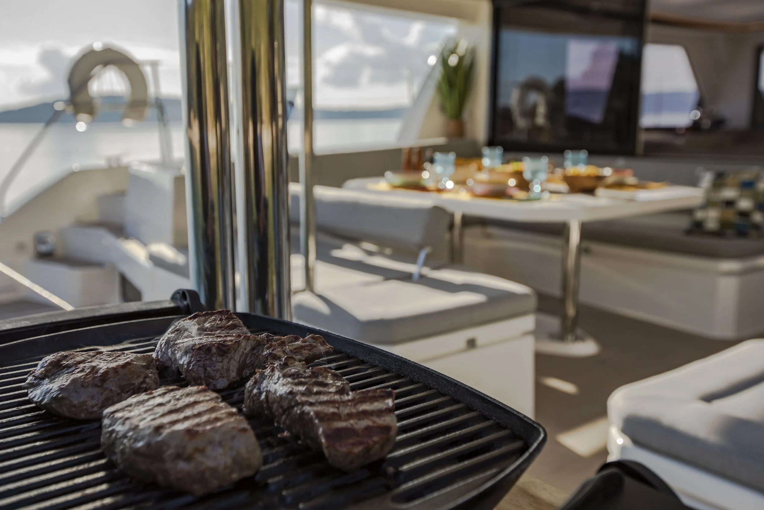 Grilling steaks on an outdoor barbecue on a boat with a dining area in the background.
