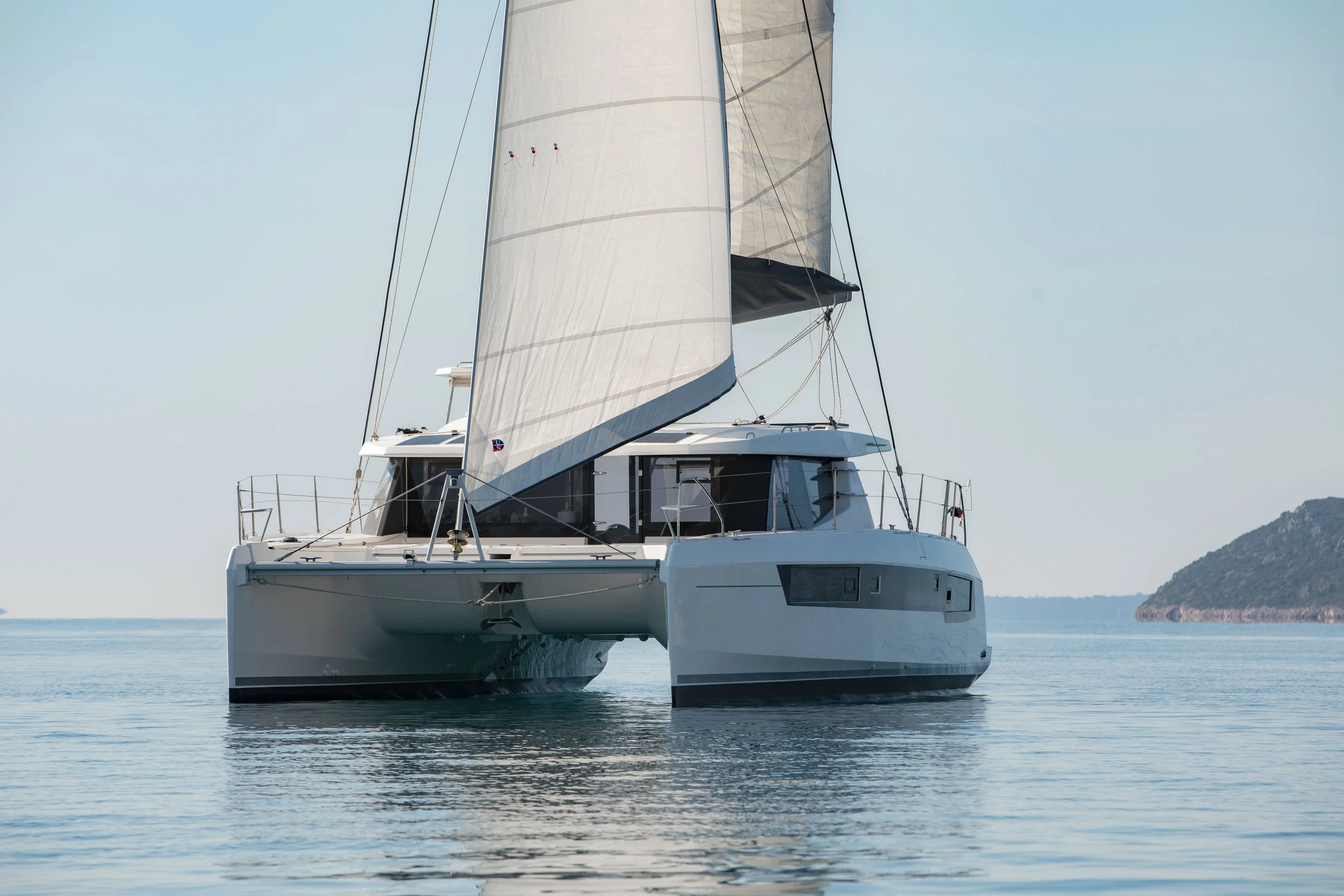 A white sailboat with a large sail on calm water, with land visible in the distance under a sky with some clouds.