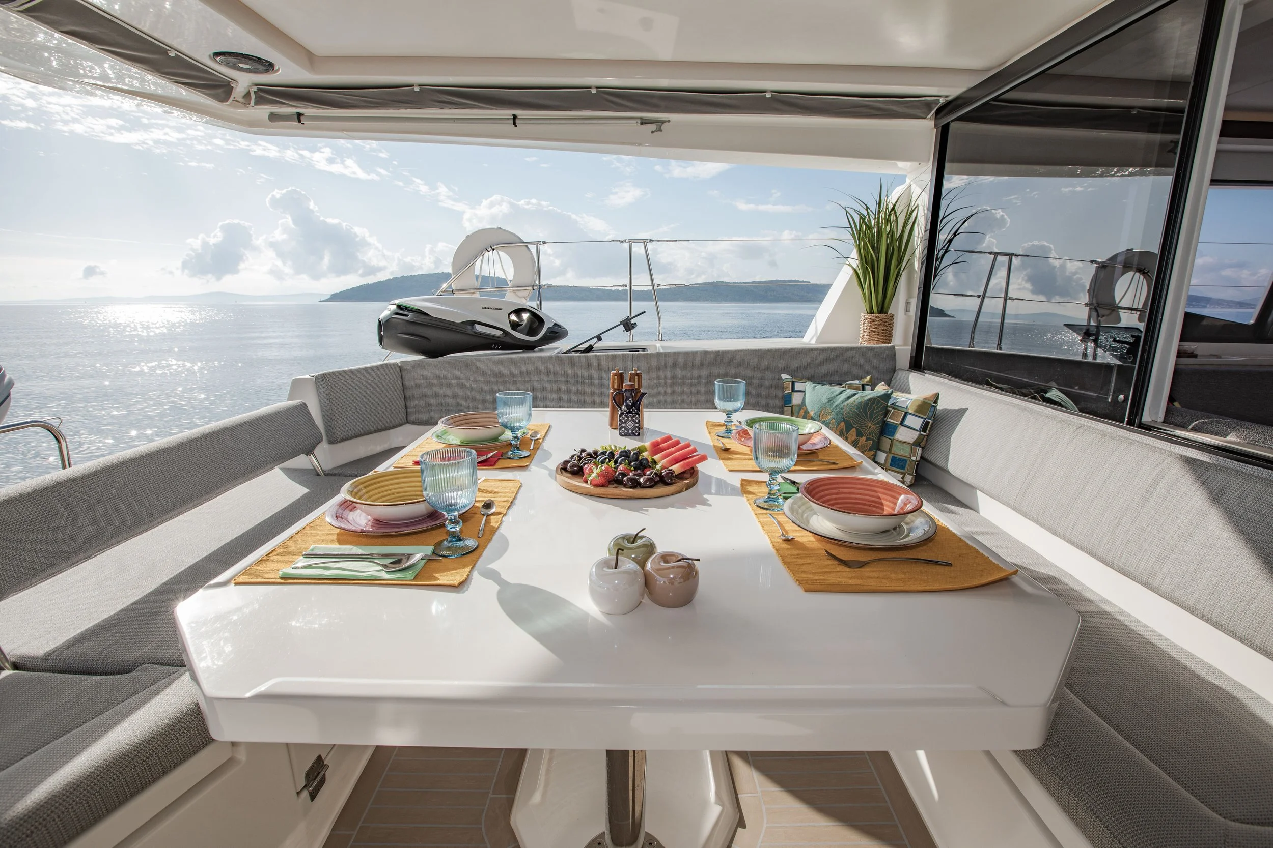 A dining setup on a yacht's outdoor deck with a table set with colorful dishes, fruit, and glasses overlooking the water and sky.