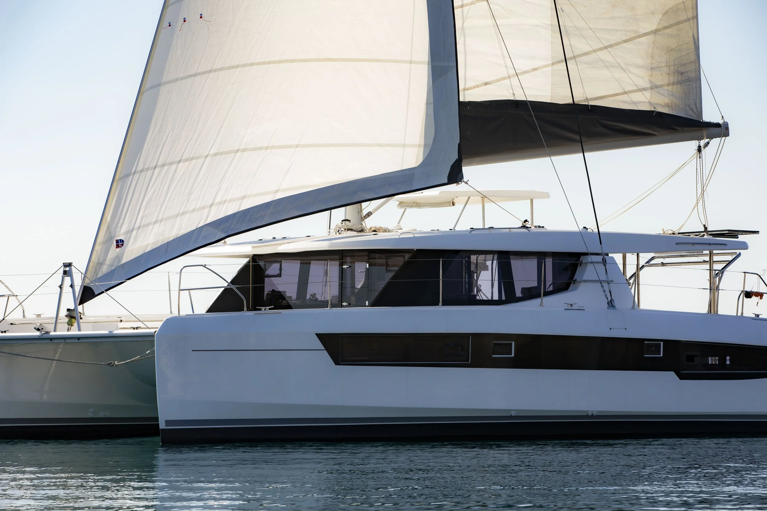 A modern white sailboat with a large sail on the water, with another boat partially visible beside it.