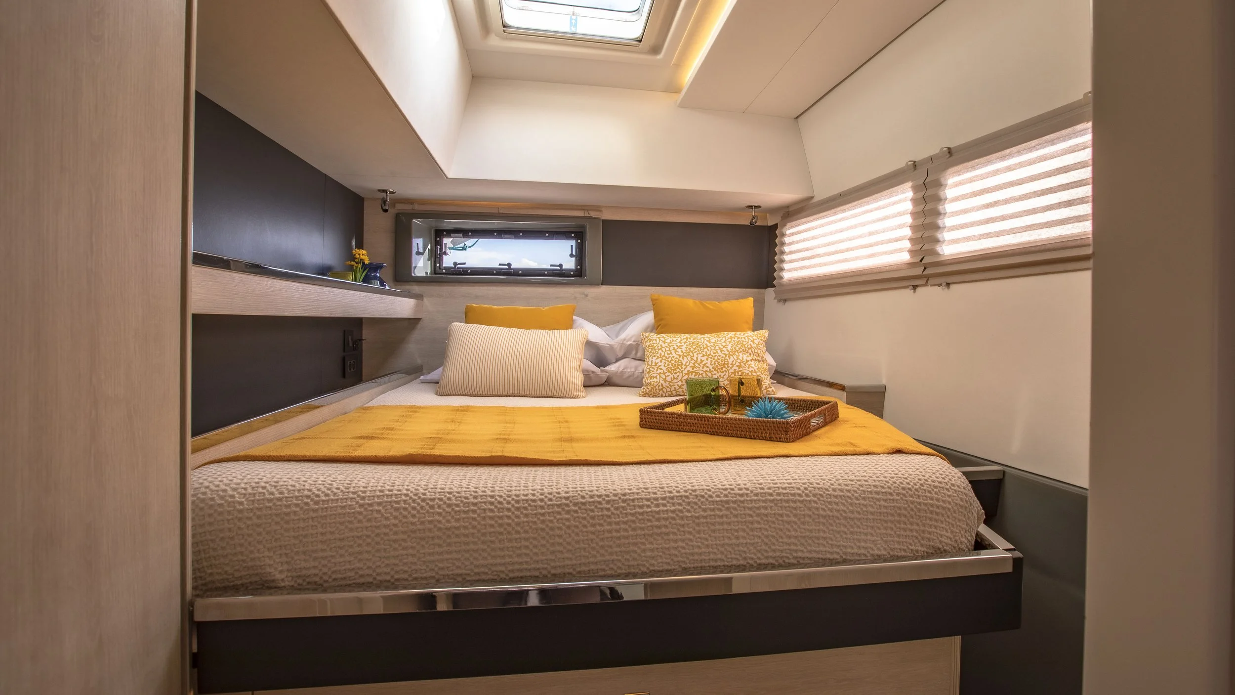 A cozy bedroom with a bed dressed in yellow and white linens, multiple pillows, a small tray with decorative items, small windows with blinds, and a skylight overhead.