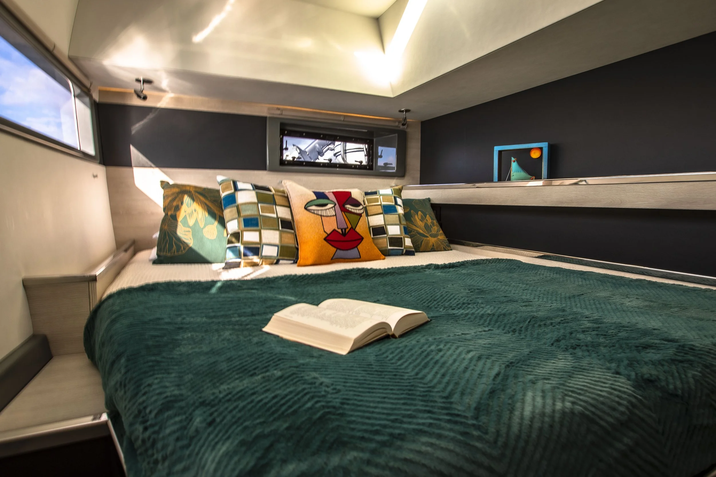 A modern bedroom with a bed covered in a teal blanket, decorated with patterned and colorful pillows, with an open book on the bed, and small windows on the wall allowing natural light to enter.