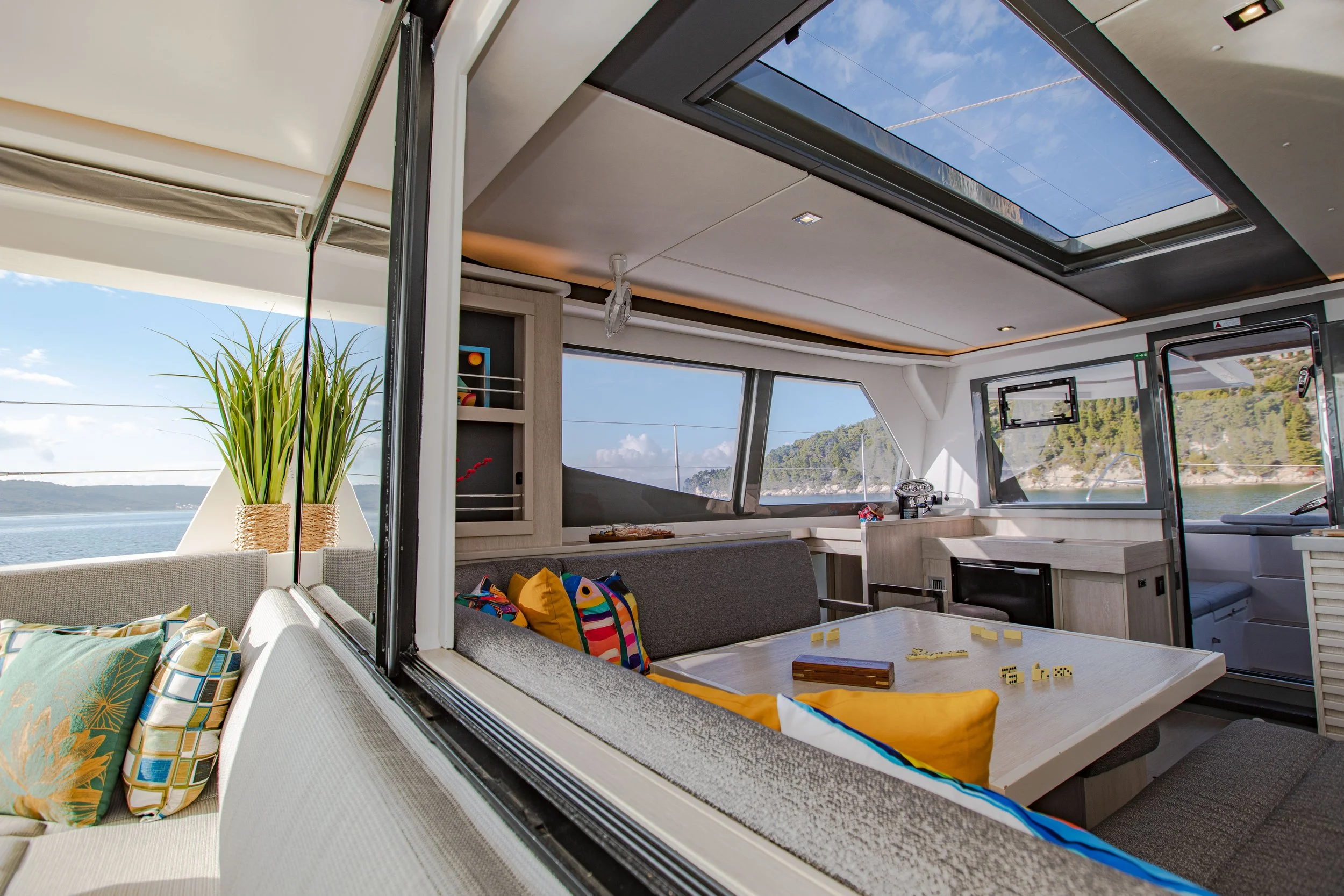 Interior of a yacht with seating area, pillows, a table with dominoes and dice, large windows showing water and landscape outside, and a ceiling skylight.