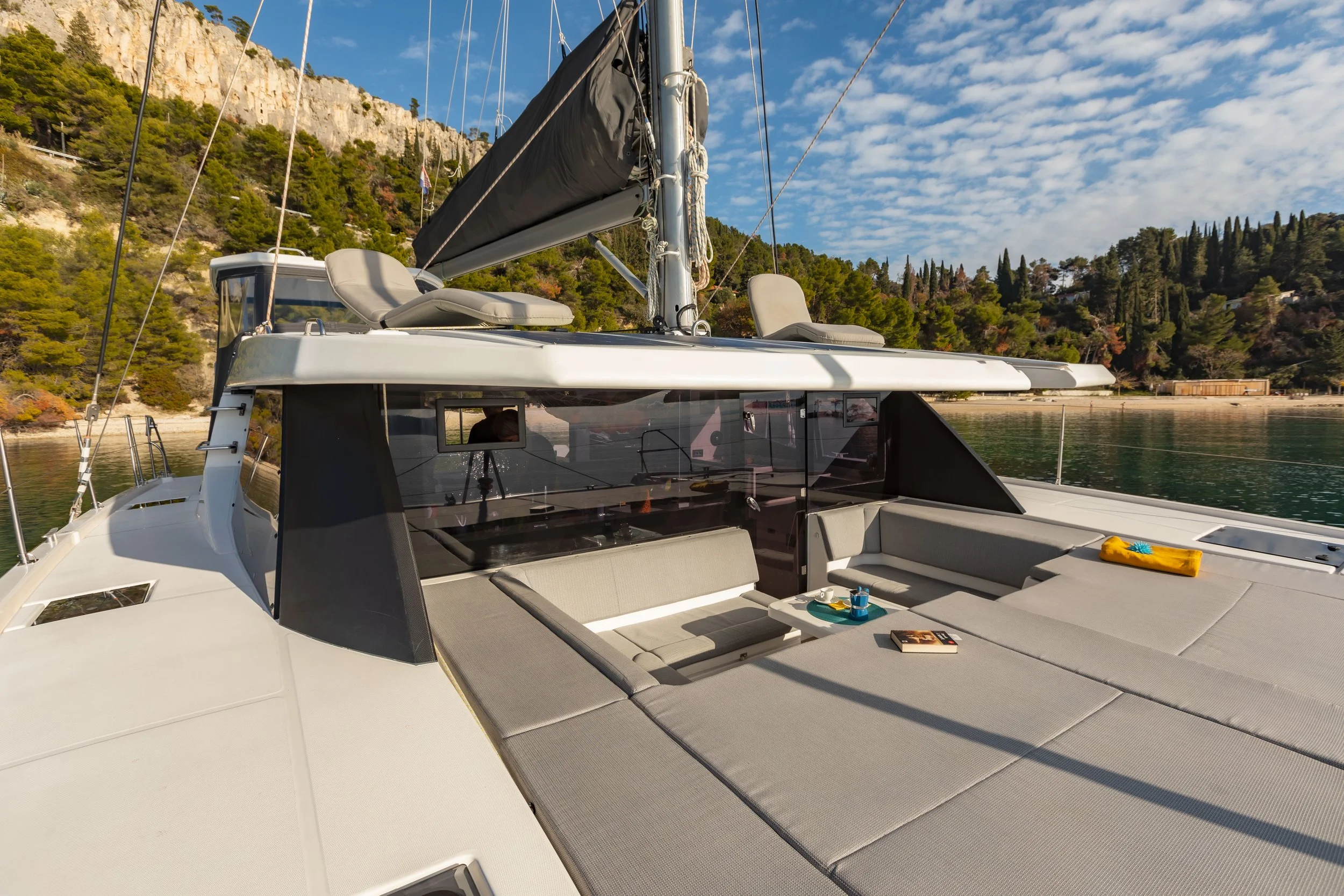 A luxury yacht anchored near a beach with a rocky shoreline and wooded hills in the background, featuring a spacious deck with cushioned seating, a table, and a partially furled sail.