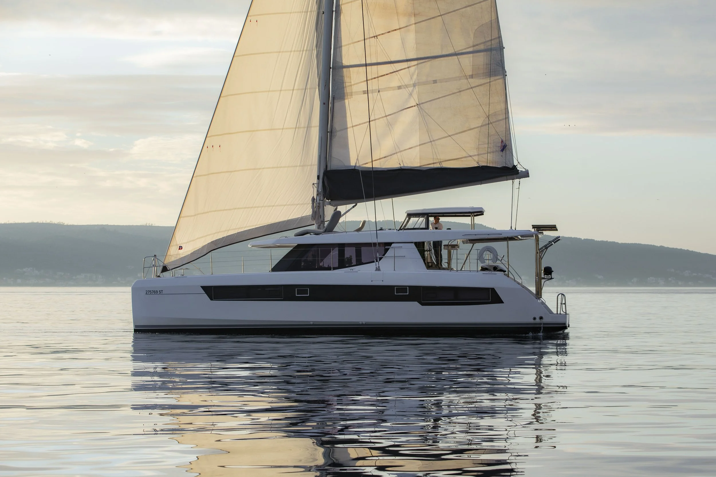 A white sailing yacht with large beige sails on calm water during sunset, with hills in the background.