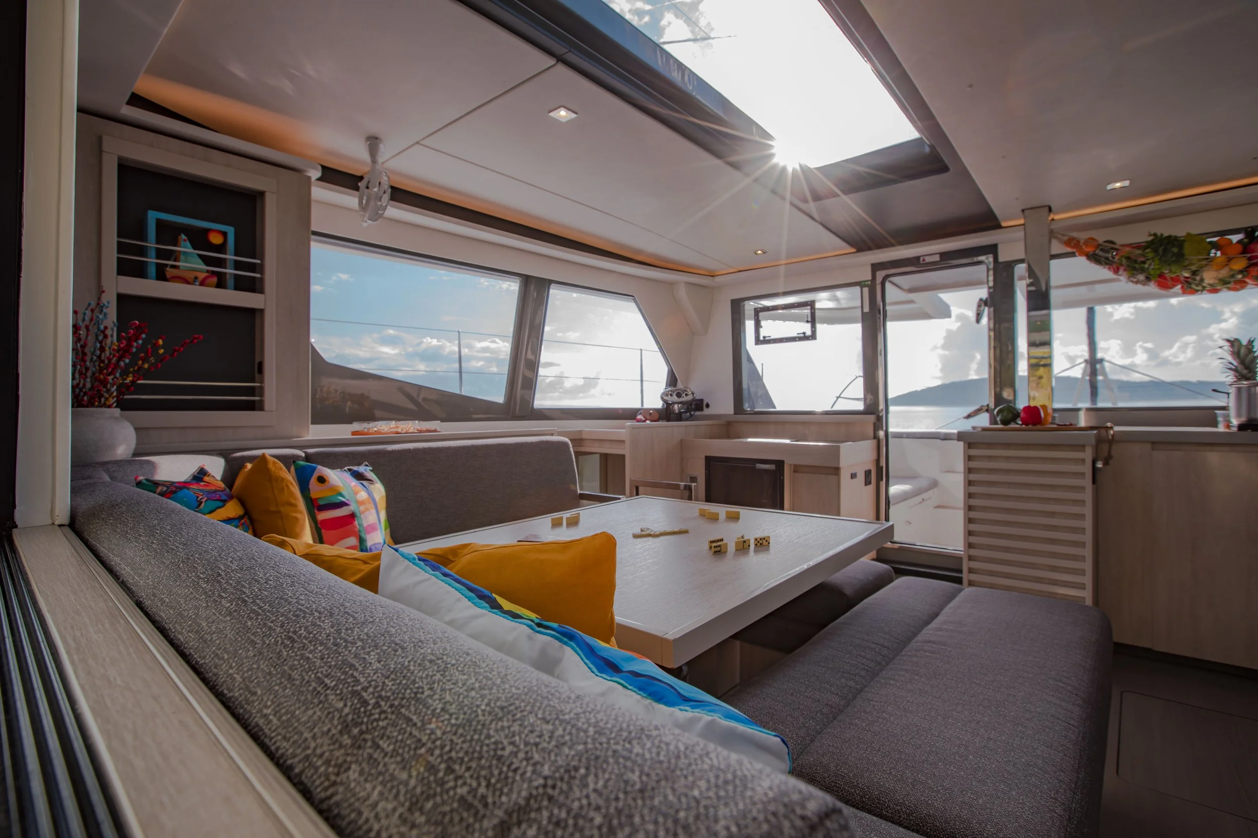 Inside a yacht with a dining area featuring a table with colorful cushions, large windows showing a scenic ocean view, and decorated with potted plants and hanging flowers.