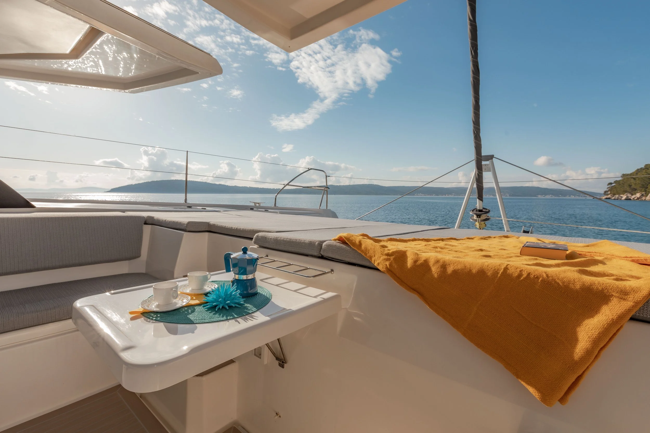 On a yacht with a view of the water and distant hills, a white table with cups and a blue coffee maker, cushioned seating, a large orange towel, and a box of chocolates.