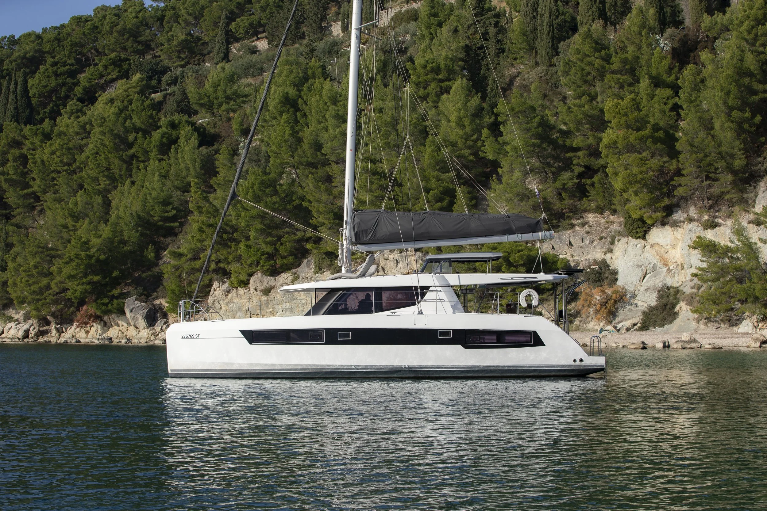 A white sailboat with a black stripe on the side is anchored in a calm body of water near a rocky shoreline covered with green pine trees.
