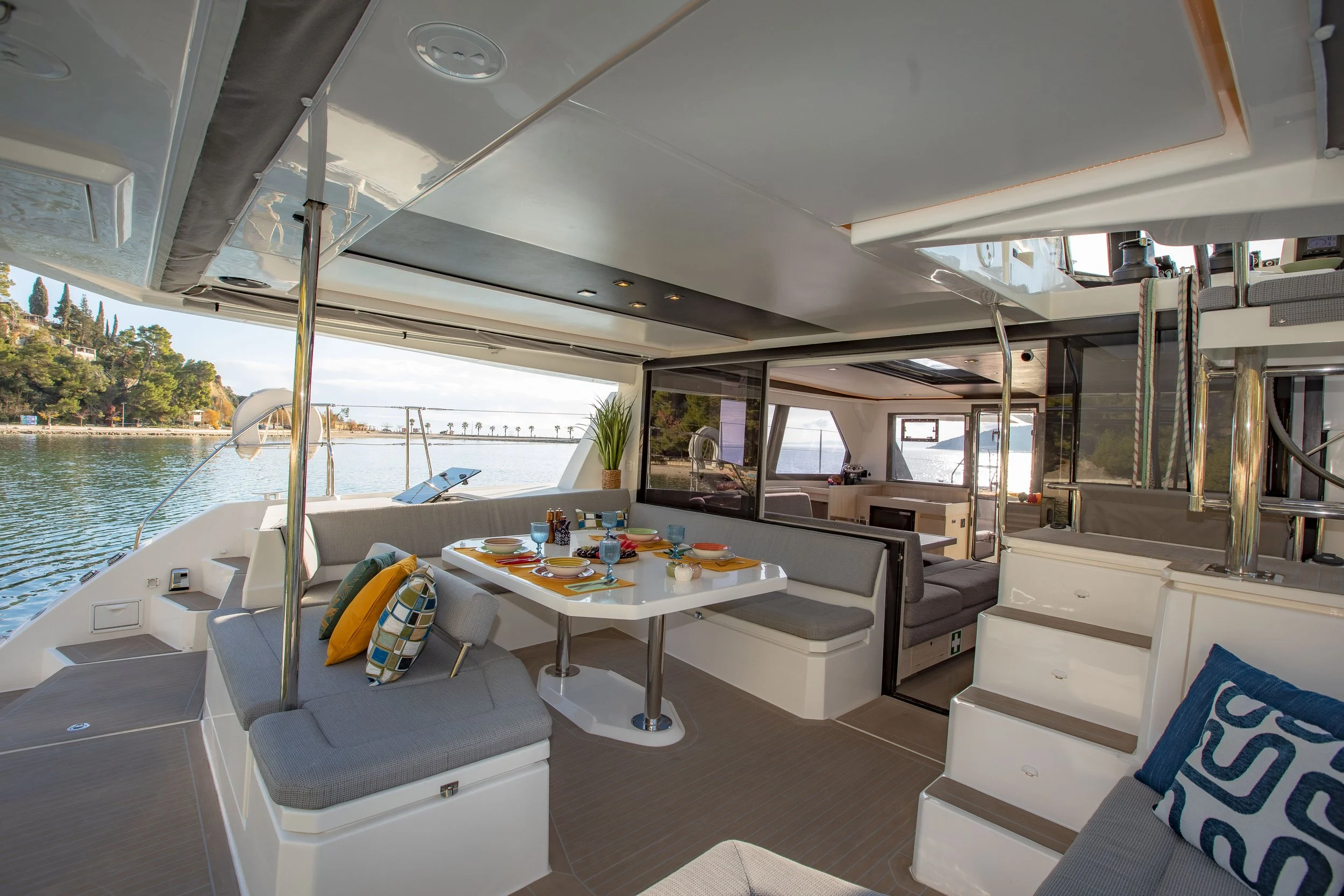 Interior of a boat with sunlight streaming in, featuring a dining table set with colorful plates, glasses, and napkins, surrounded by cushioned seating and stairs leading to an upper deck, with water and shoreline visible outside.
