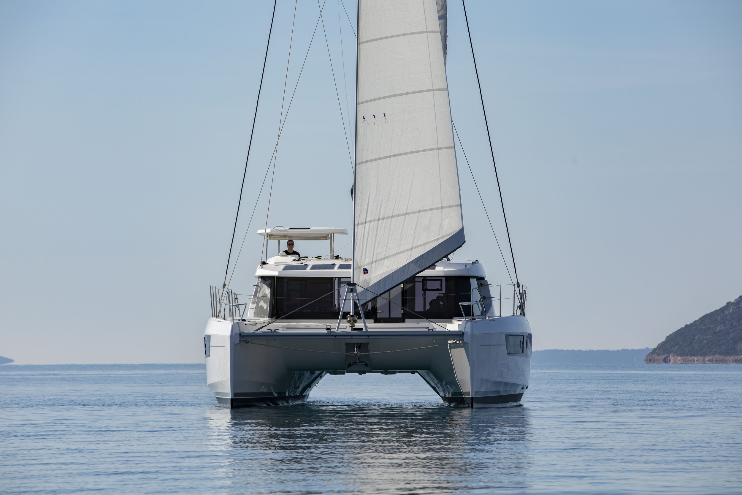 A modern white sailboat on calm water with a person on deck, with distant landmasses and a clear blue sky in the background.
