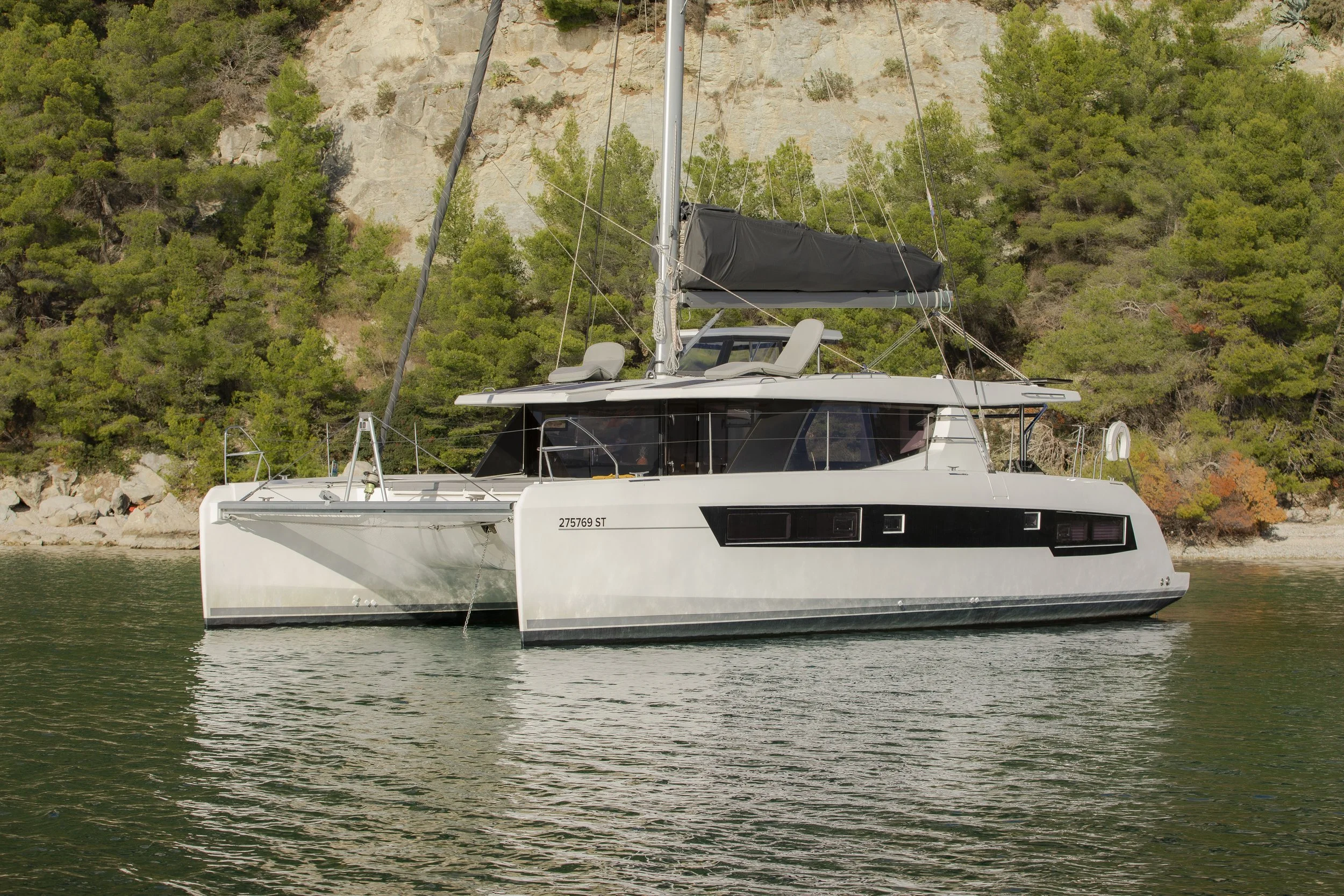 A modern sailboat with a white hull and black accents floating on calm water near a rocky shoreline and green trees.