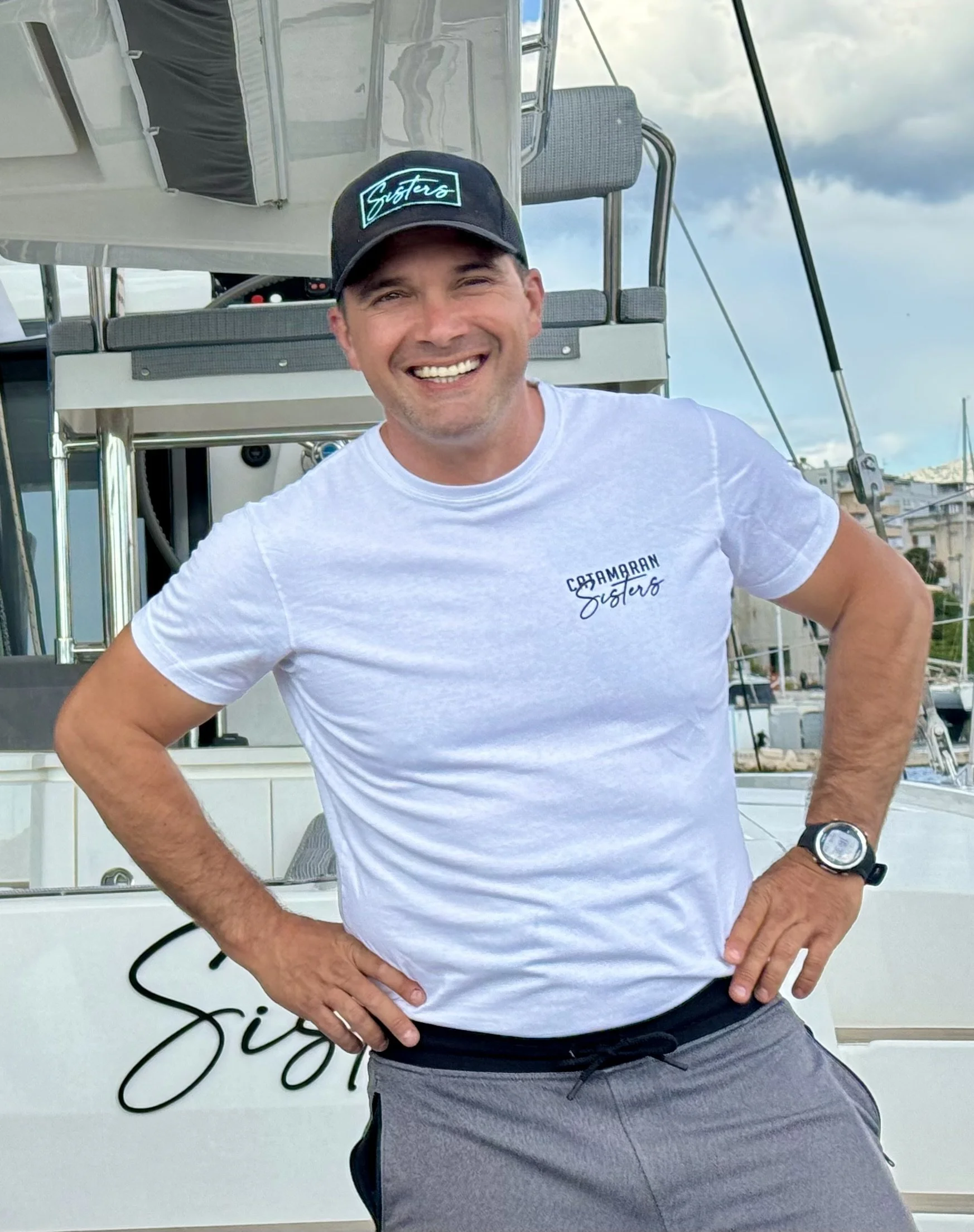 A smiling man in a white t-shirt and gray shorts standing on a boat with hands on hips, wearing a black watch and a black cap, with a marina and cloudy sky in the background.